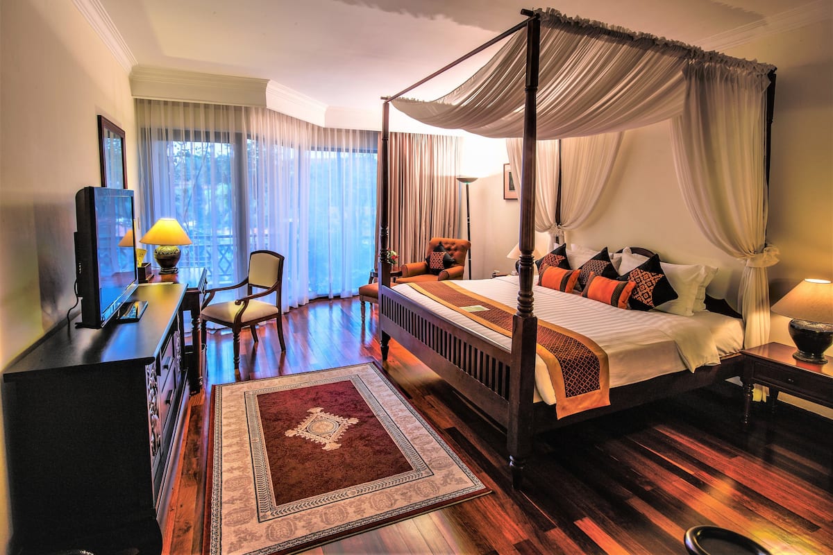 A four-poster canopy bed with a wooden frame, a red and beige patterned area rug, a wooden desk with a lamp, and a television on a dresser.