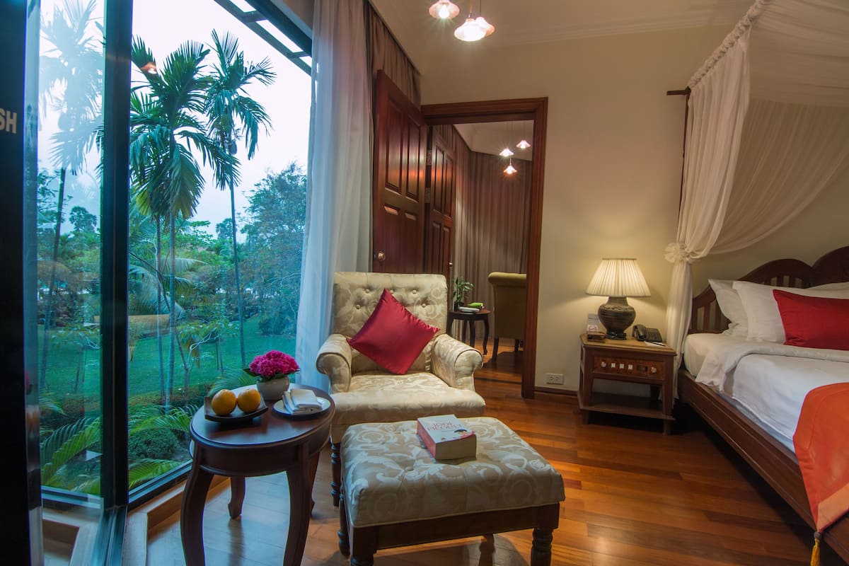 A hotel room with a bed, a chair, a table with fruit and flowers, a nightstand with a lamp, and a view of palm trees through large windows.