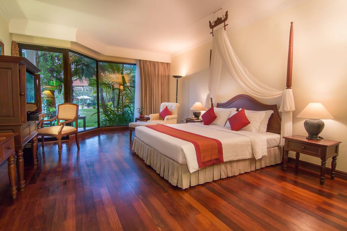 A spacious bedroom with a large bed, a canopy, and a view of a tropical garden.