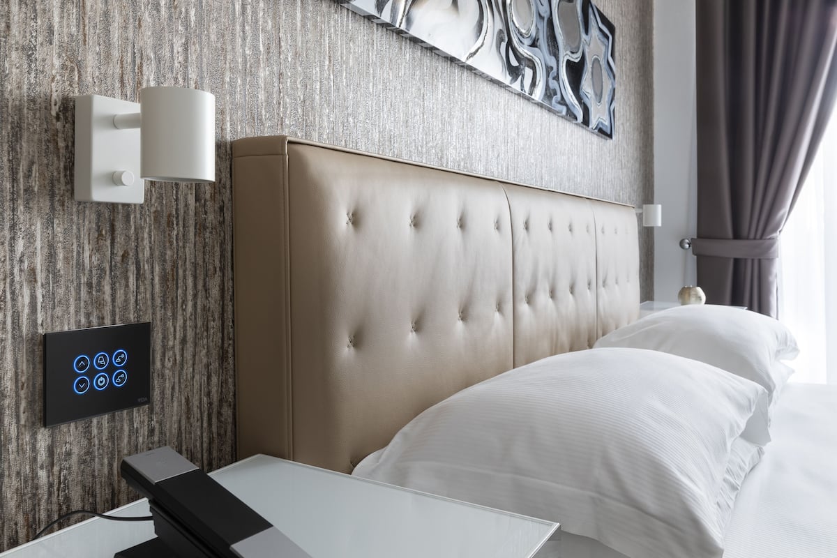 Une chambre d’hôtel moderne avec une tête de lit capitonnée, une table de chevet, un panneau de commande fixé au mur et une grande œuvre d’art abstraite au-dessus du lit.