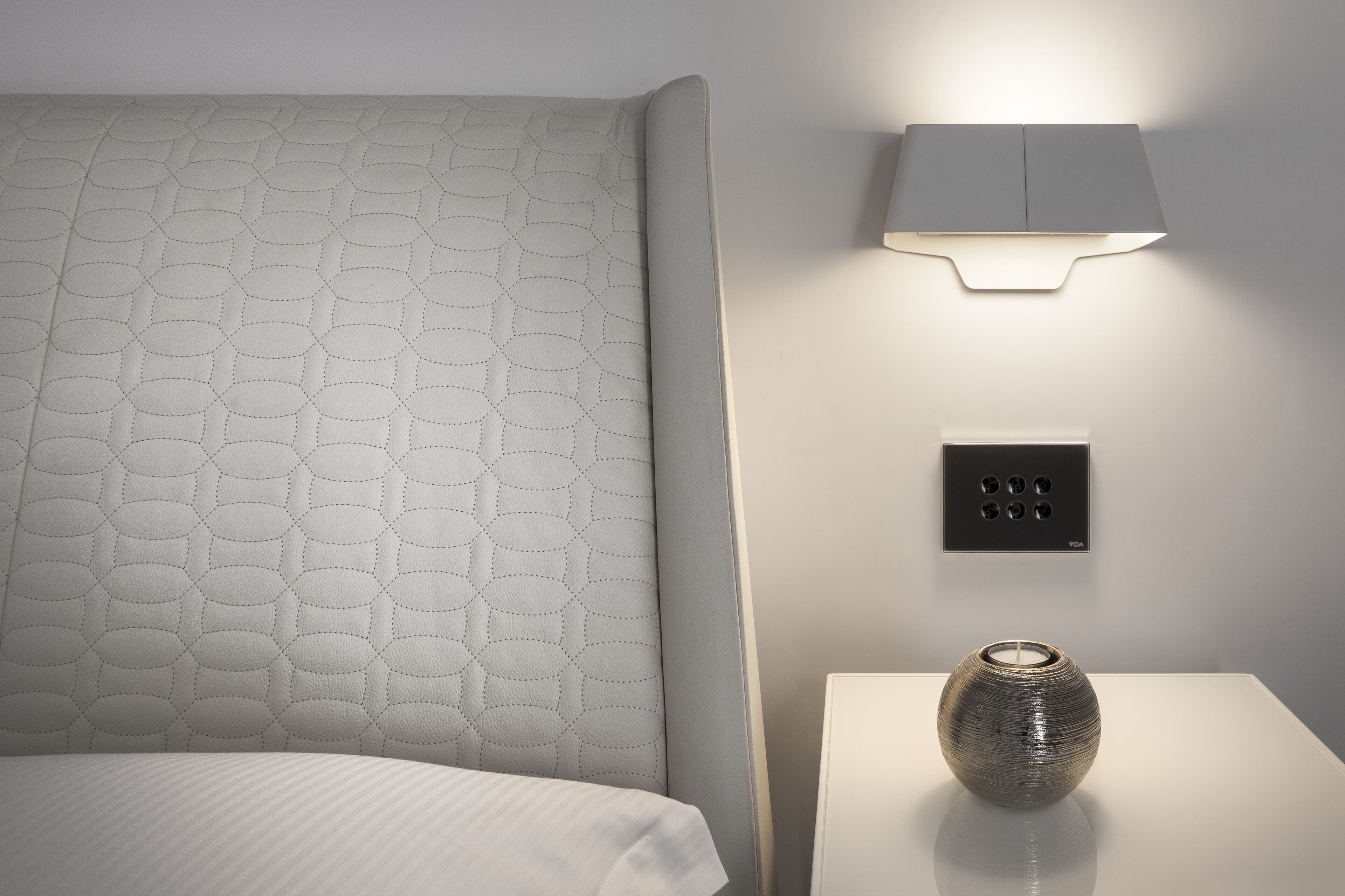 Un lit bien fait, doté d’une tête de lit texturée, d’une lampe fixée au mur, d’une prise de courant et d’un vase posé sur une table de chevet.