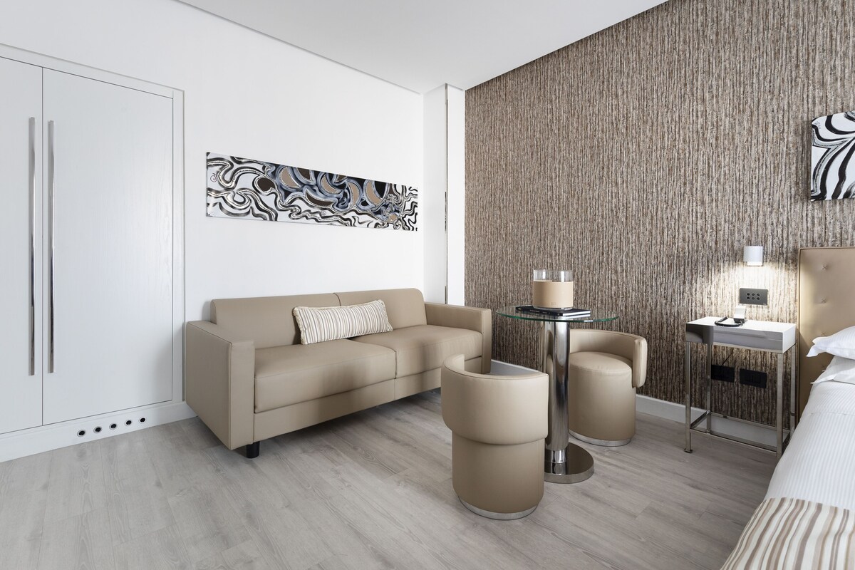 Ein modernes Hotelzimmer mit beiger Couch, rundem Couchtisch, einem kleinen Beistelltisch und einem Bett mit gestreifter Bettwäsche.