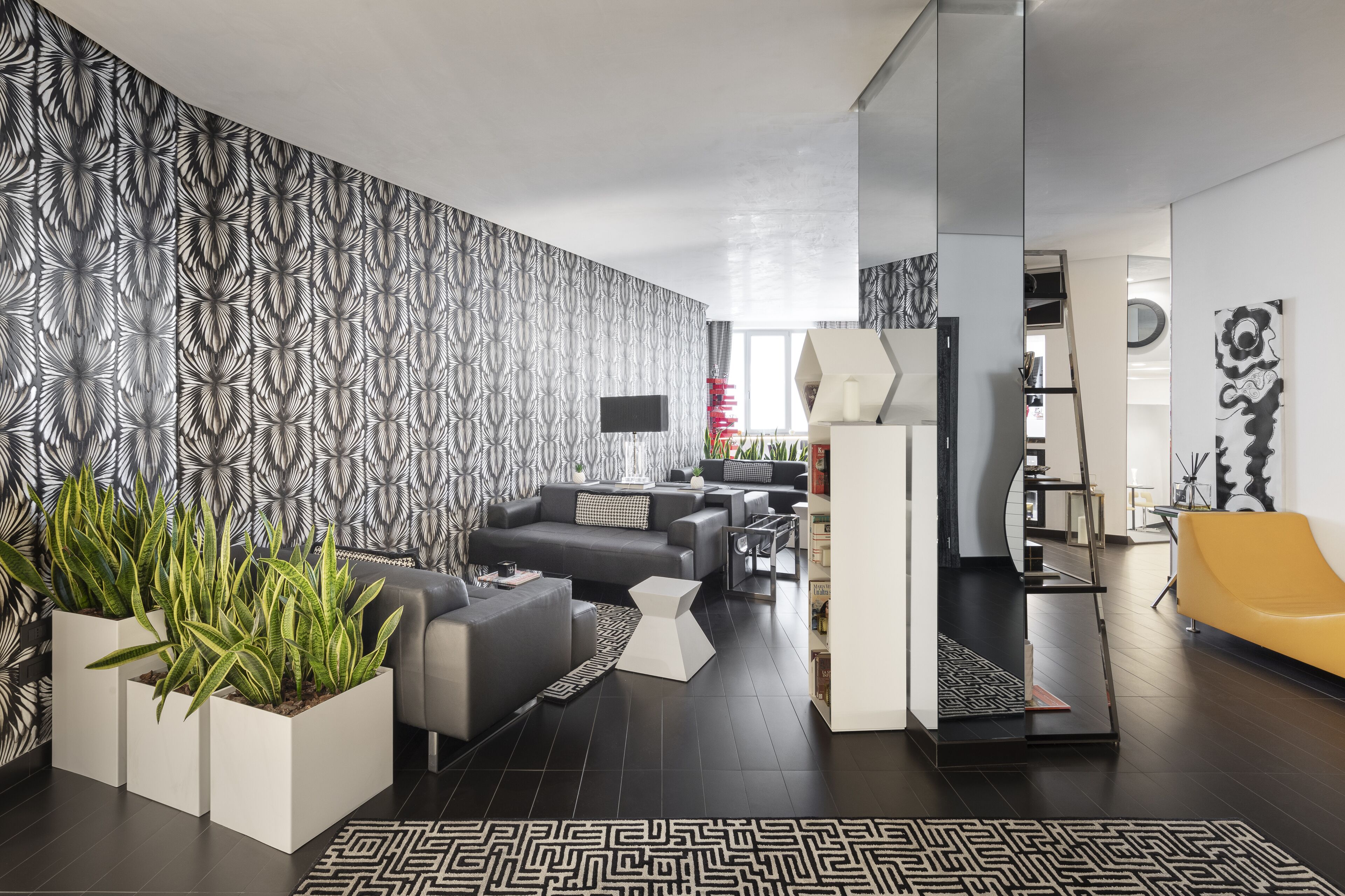Una sala de estar moderna con un sofá gris, una mesa de centro blanca y un sillón amarillo. Hay plantas en macetas y una alfombra estampada.