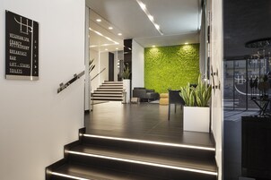 Una hall moderna di un hotel con una scala che conduce ai piani superiori, una parete verde con texture e un cartello che indica servizi come un ristorante e un bar.