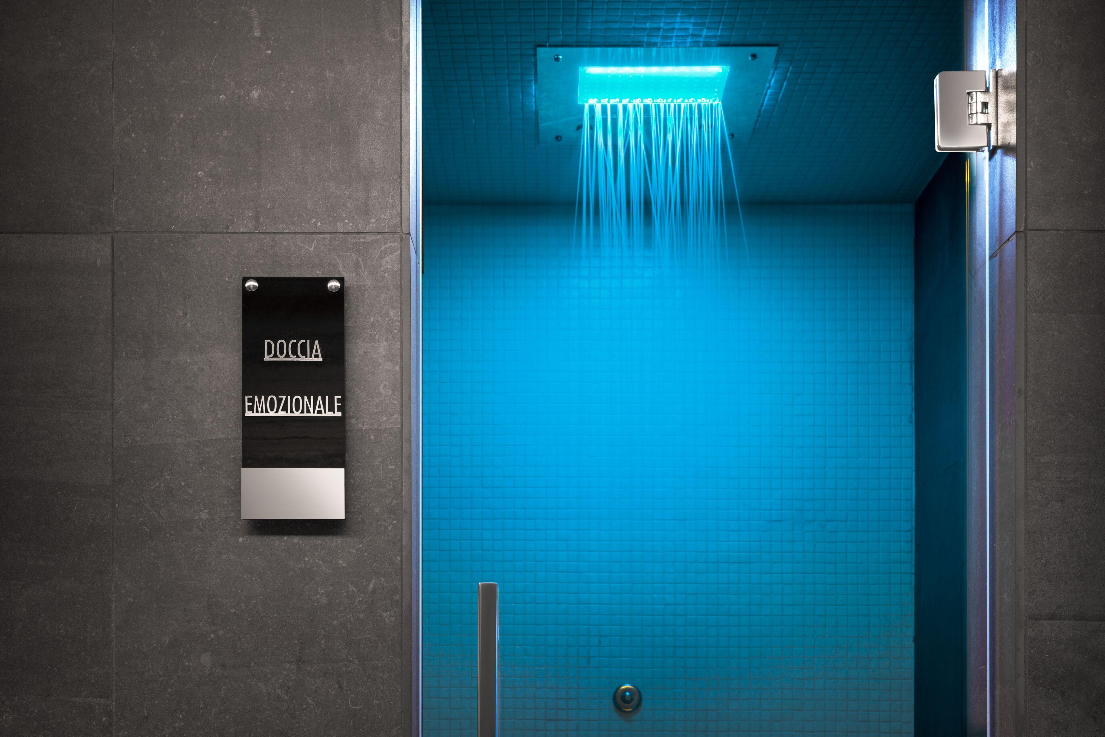 A modern bathroom with a blue tiled wall, a showerhead, and a sign that reads "DOCCIA EMOZIONALE".