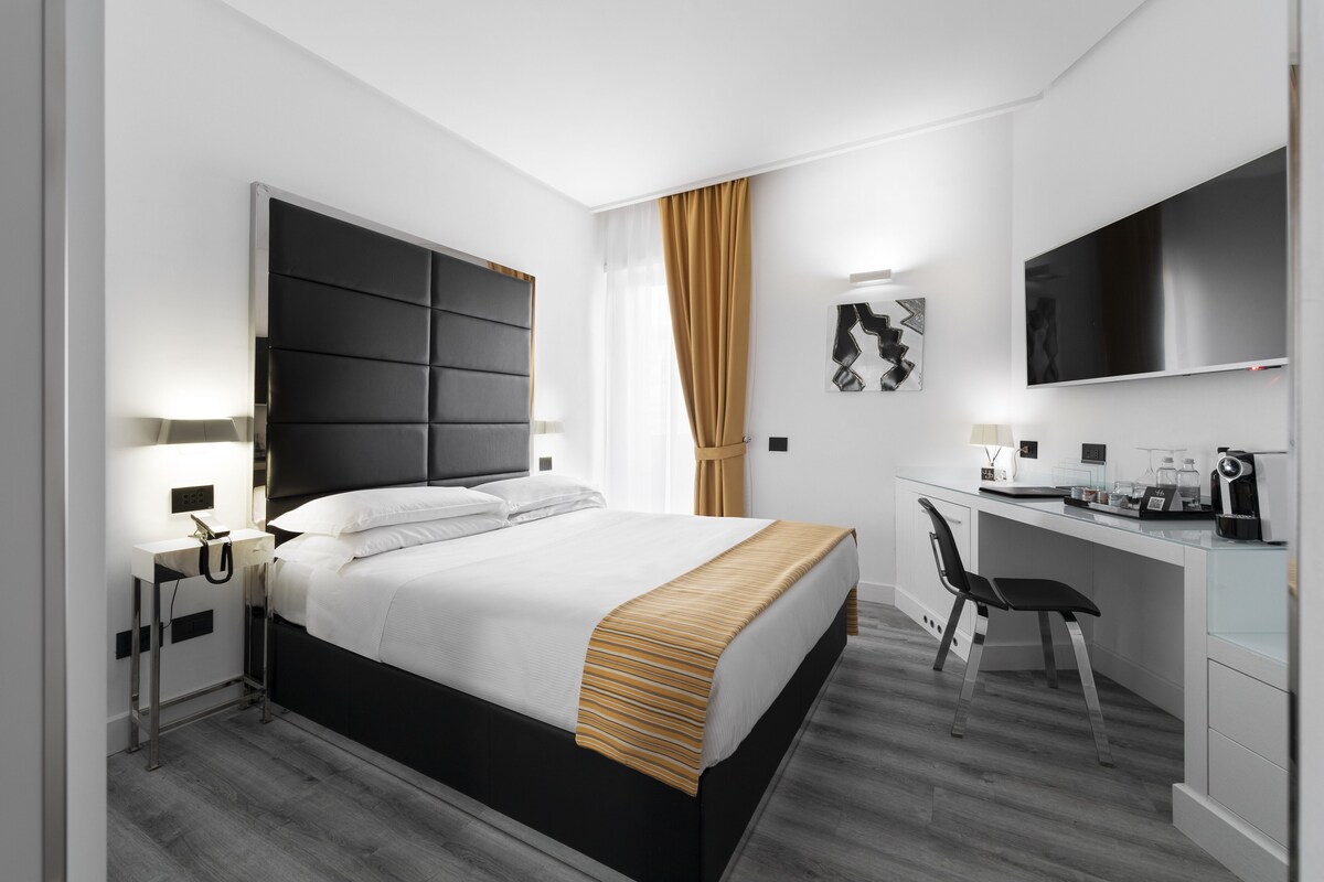 Ein modernes Hotelzimmer mit einem großen Bett, einem Schreibtisch mit Stuhl, einem Flachbildfernseher und einer kleinen Küchenzeile.