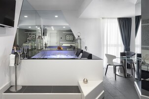 Una moderna camera d'albergo con un ampio specchio che riflette l'interno della stanza, un tavolo con piano in vetro e sedie, un piccolo angolo salotto e un bagno con vasca da bagno ben visibile.