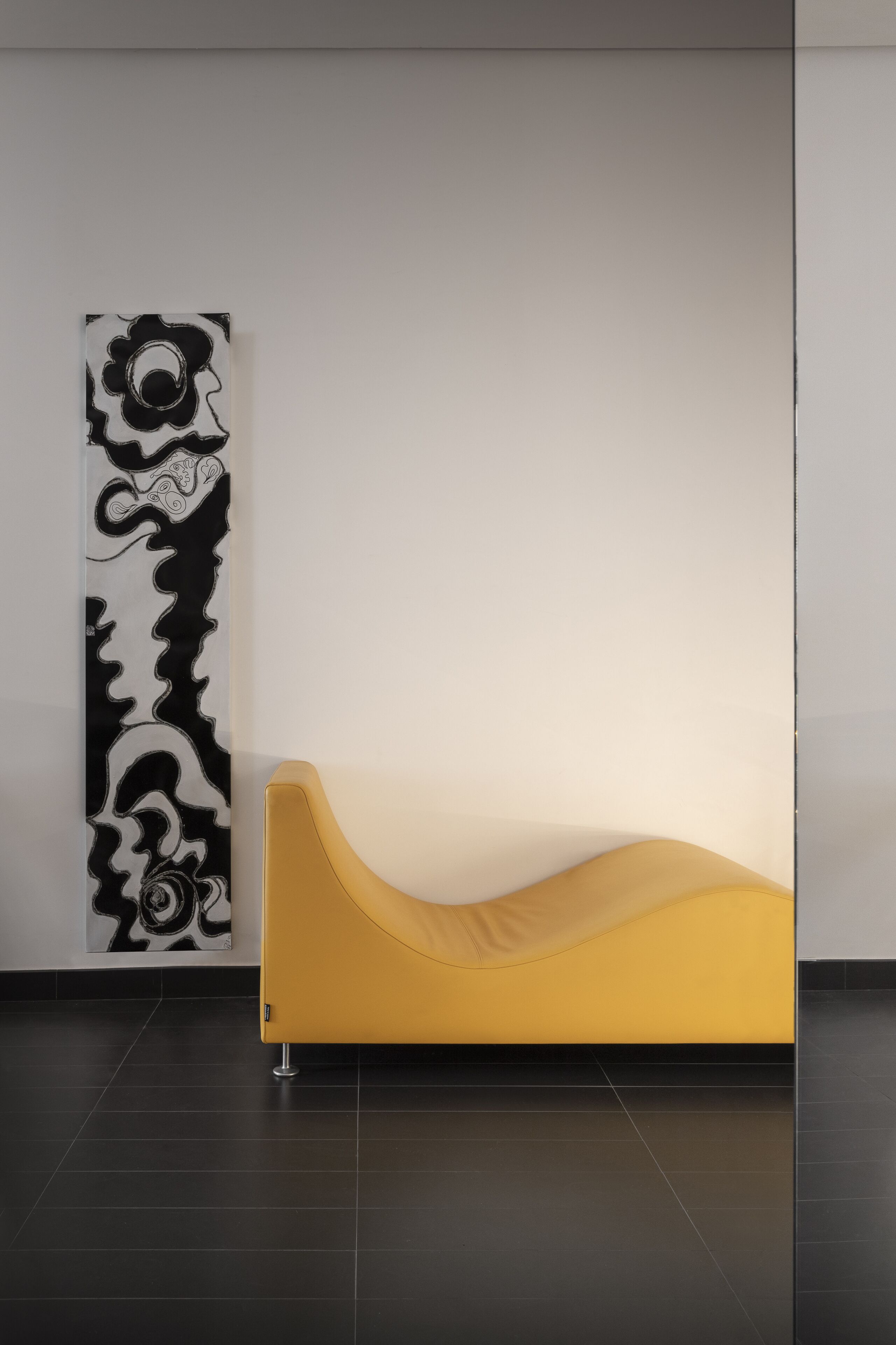 Un interior moderno con un sofá amarillo frente a una pared blanca, un cuadro abstracto en blanco y negro y un piso de baldosas oscuras.