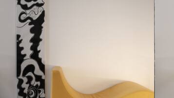 Un interior moderno con un sofá amarillo frente a una pared blanca, un cuadro abstracto en blanco y negro y un piso de baldosas oscuras.