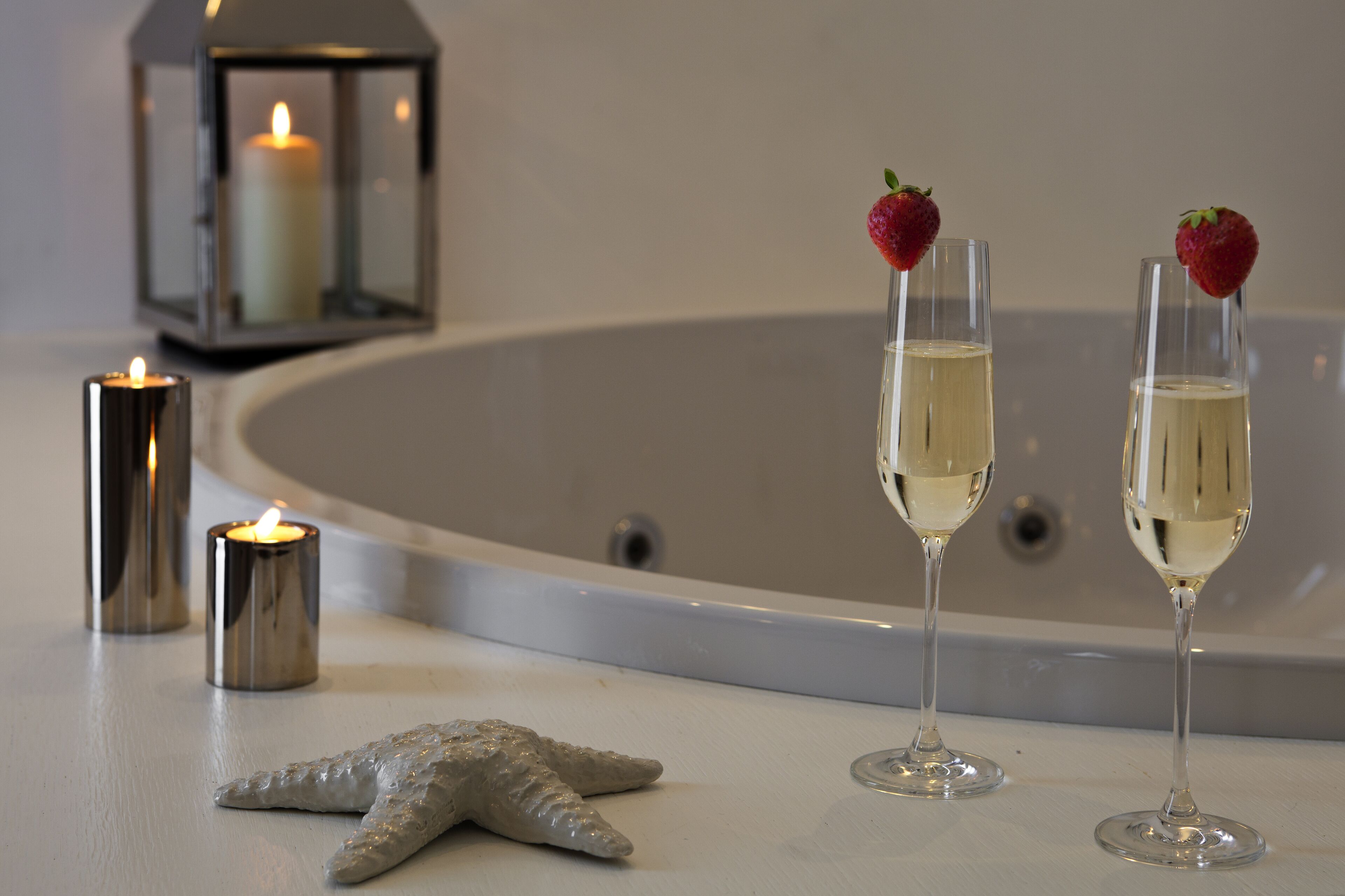 Una vasca da bagno con due bicchieri da champagne, ognuno decorato con una fragola, e una candela accesa su una superficie bianca.
