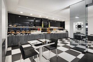 Een moderne hotelkeuken met een geruit vloertje, een buffet met diverse gerechten en een eetgedeelte met een tafel en stoelen.