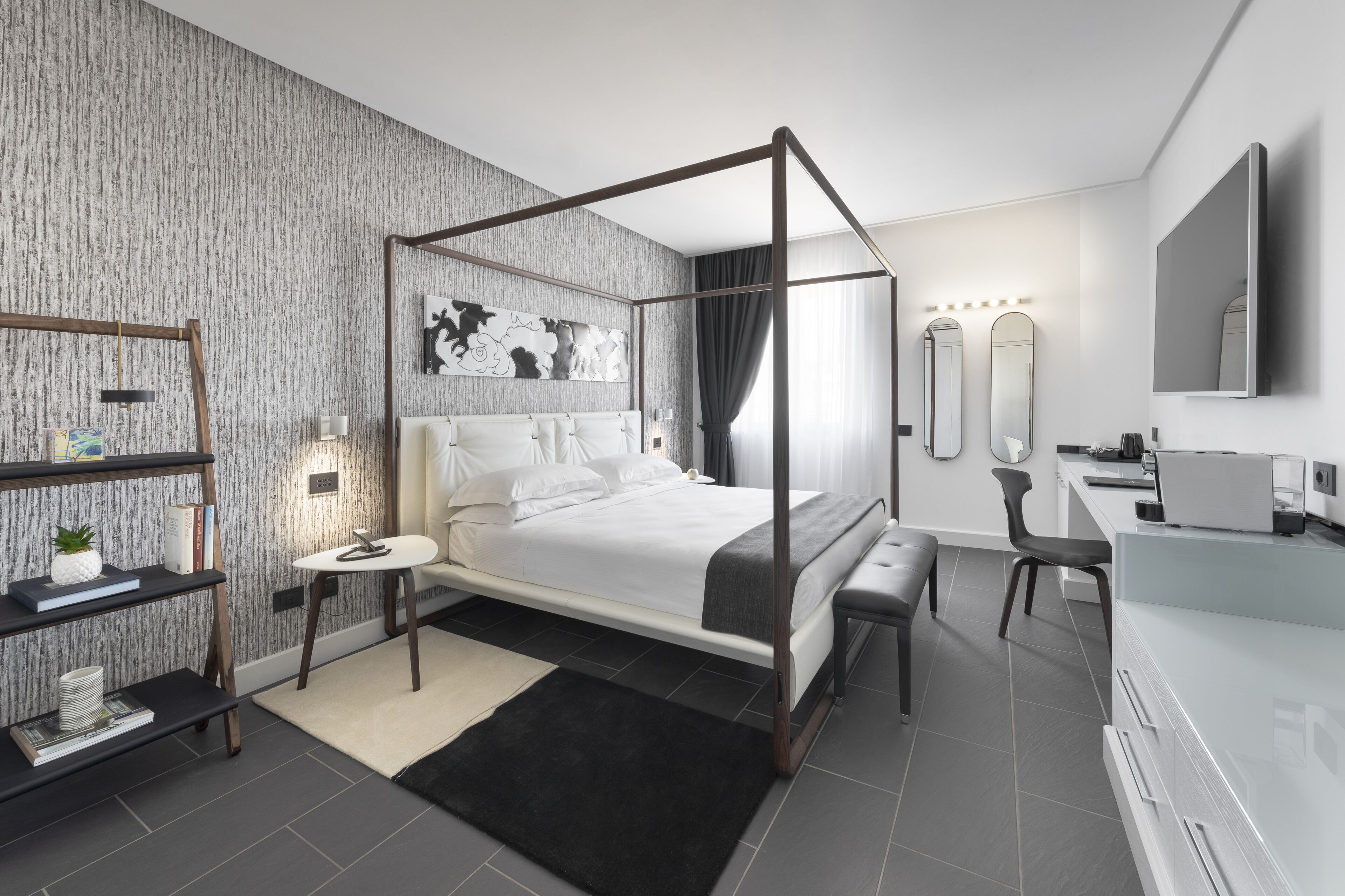 A modern bedroom with a four-poster bed, a bedside table, a desk with a chair, and a large mirror.