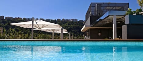 Uma piscina com superfície de água azul cristalina, um guarda-sol branco e um edifício moderno com fachada de vidro ao fundo.