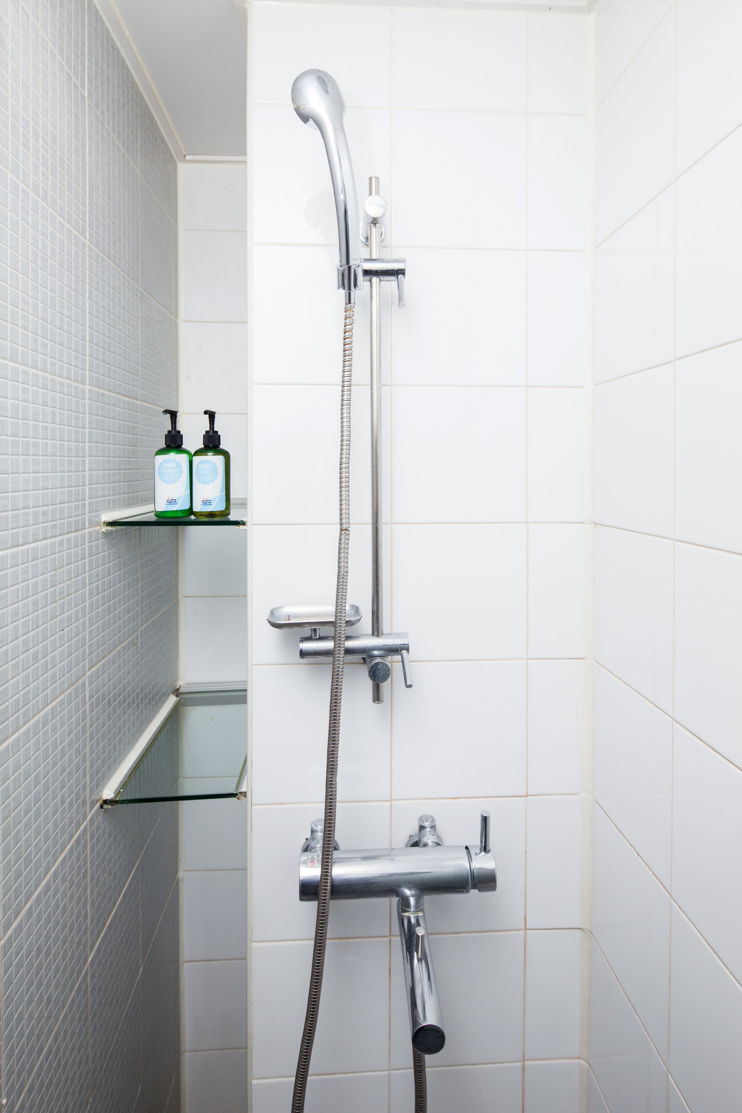Une salle de bain avec une douche, une étagère en verre où sont rangés les articles de toilette et un mur carrelé.