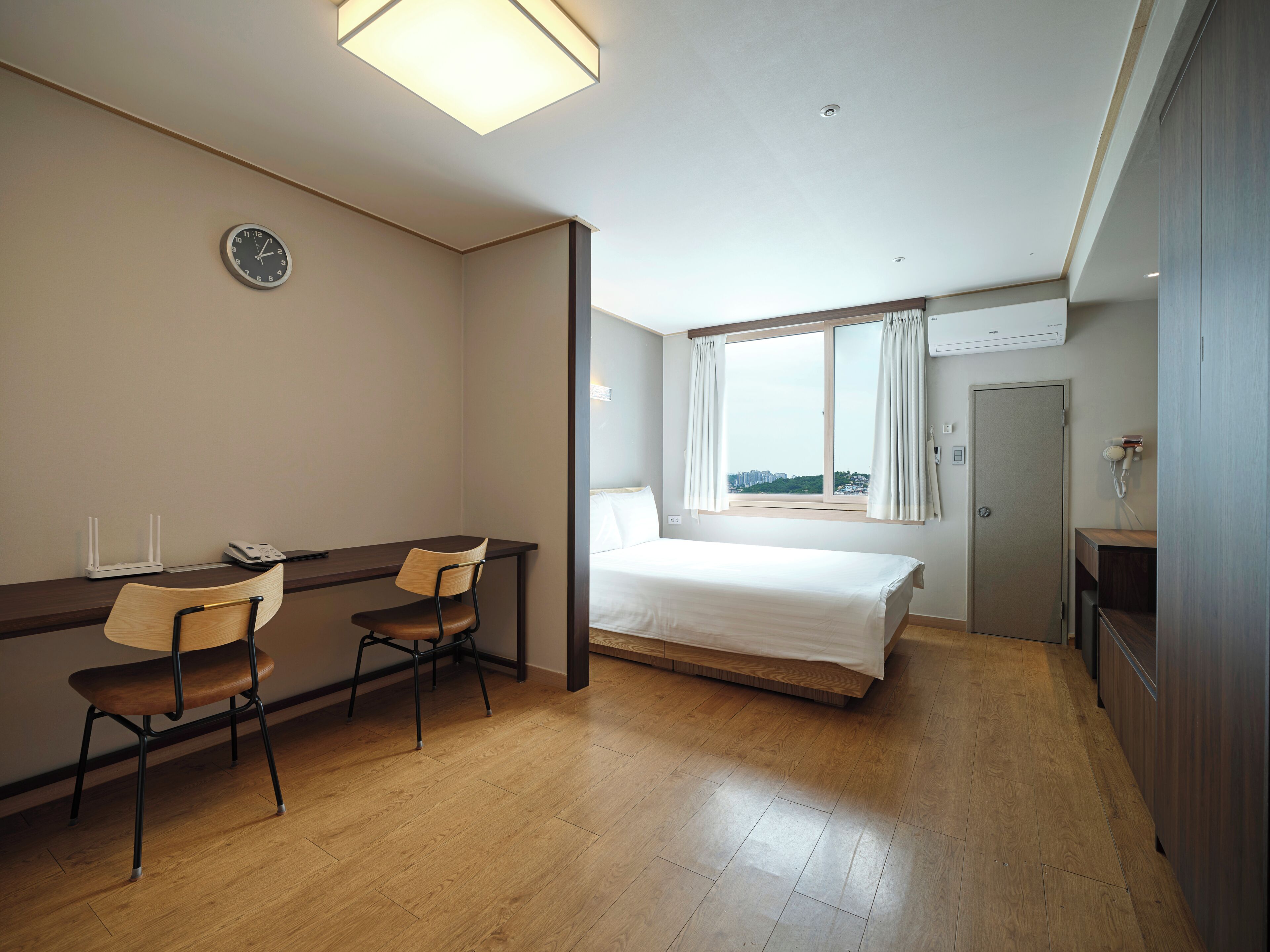Een hotelkamer met een bed, een bureau met stoelen, een klok, een raam met gordijnen en een deur.