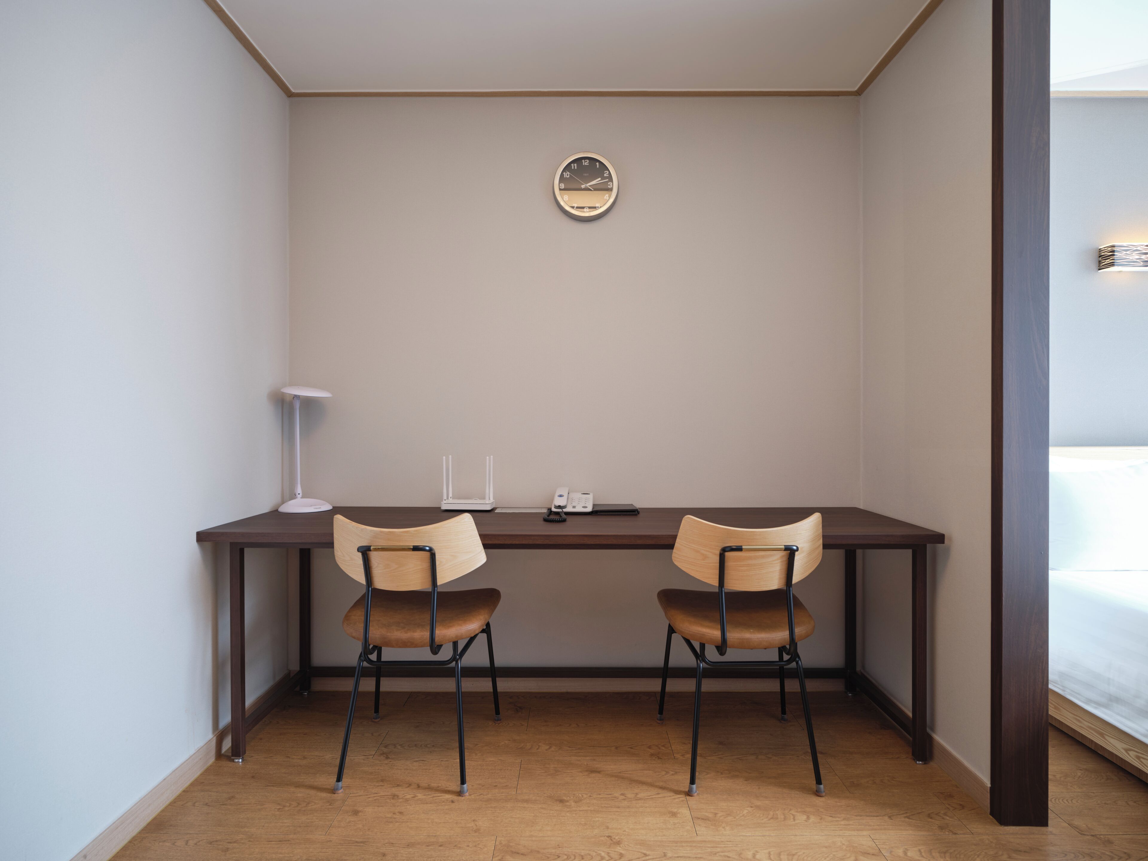 A room with a desk, two chairs, a clock on the wall, a lamp, and a bed in the corner.