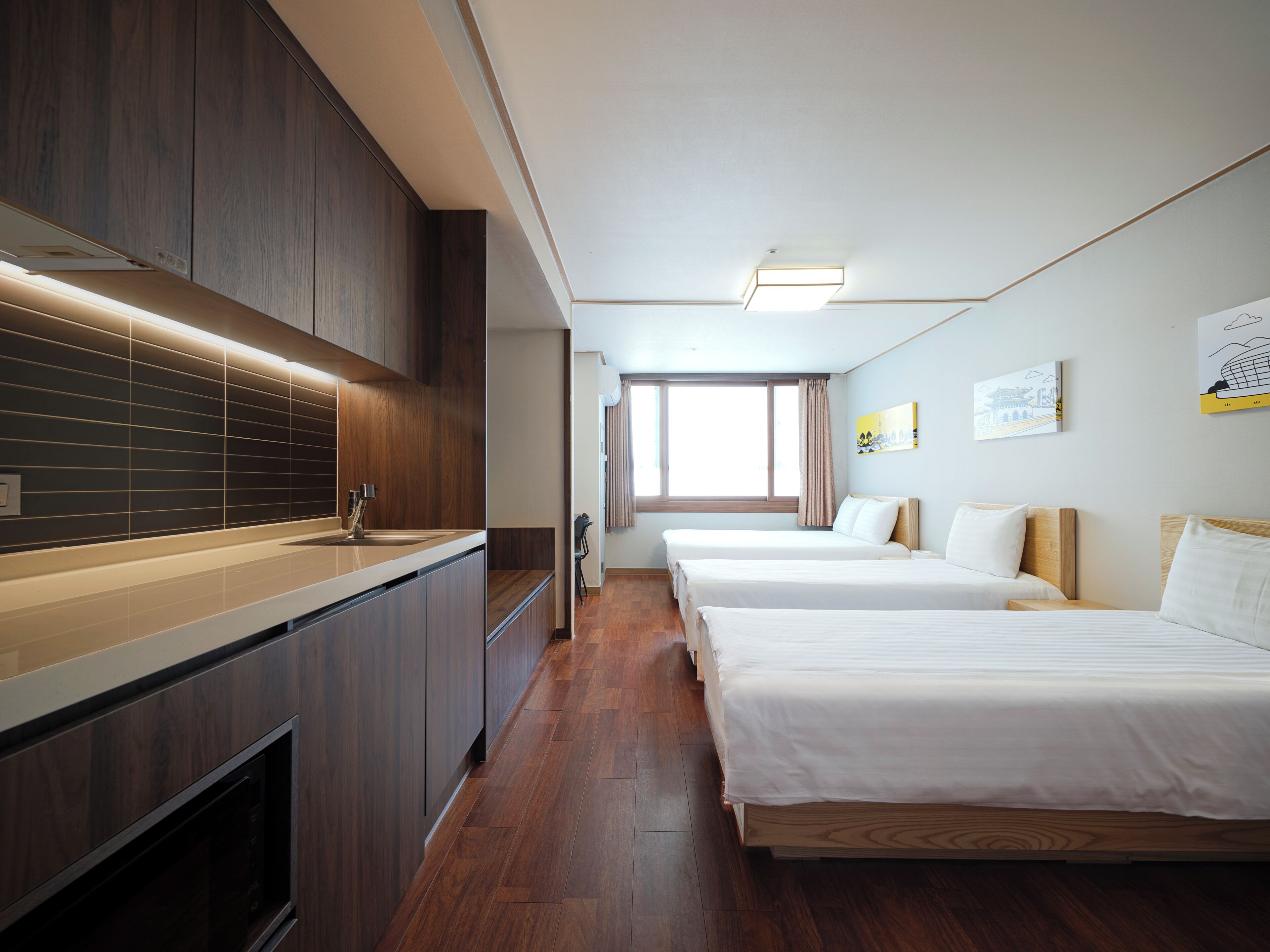 A hotel room with a kitchenette, two beds, a large window, and artwork on the walls.