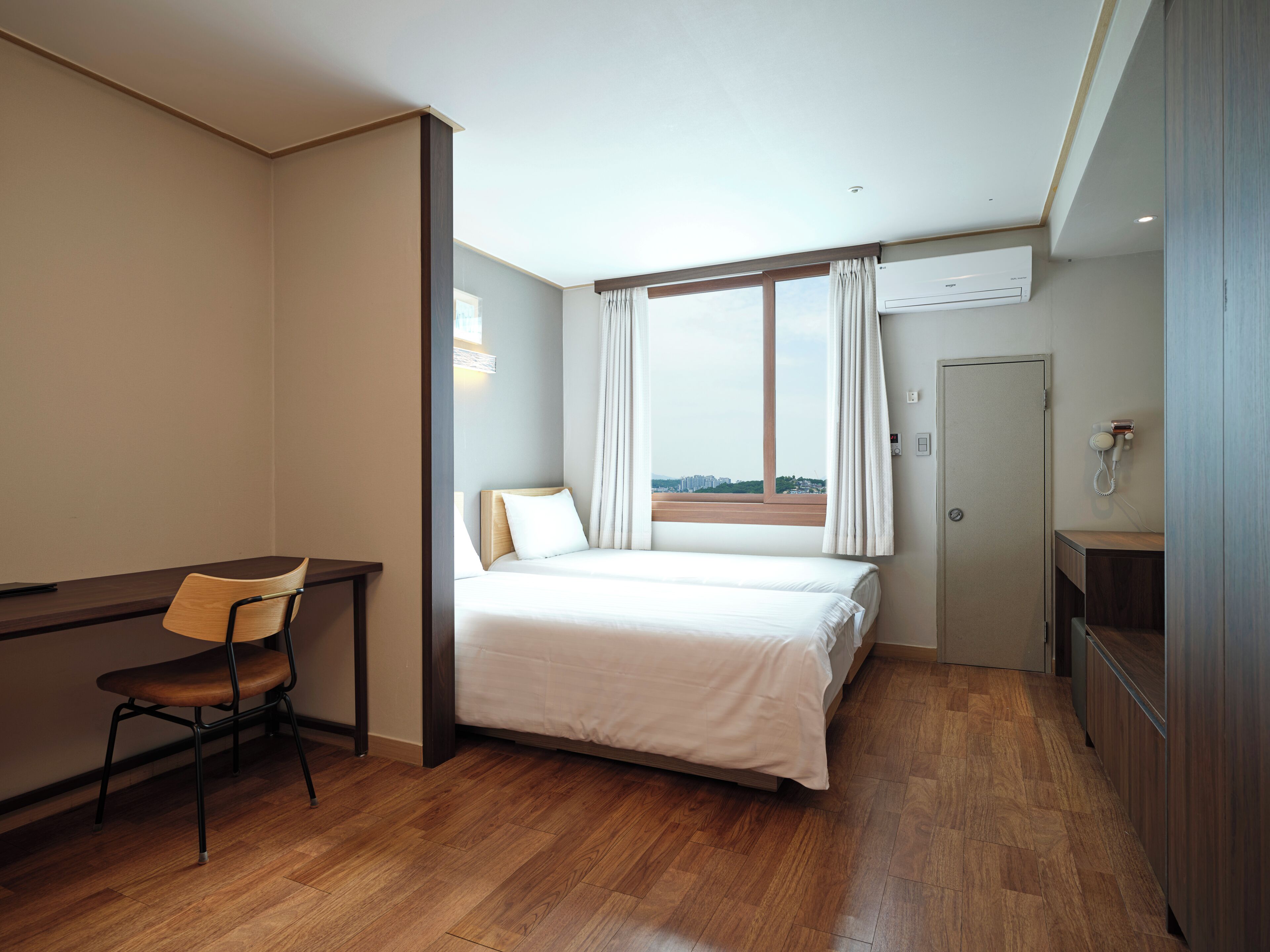 A hotel room with a bed, a desk, a chair, a window with curtains, an air conditioning unit, and a door.