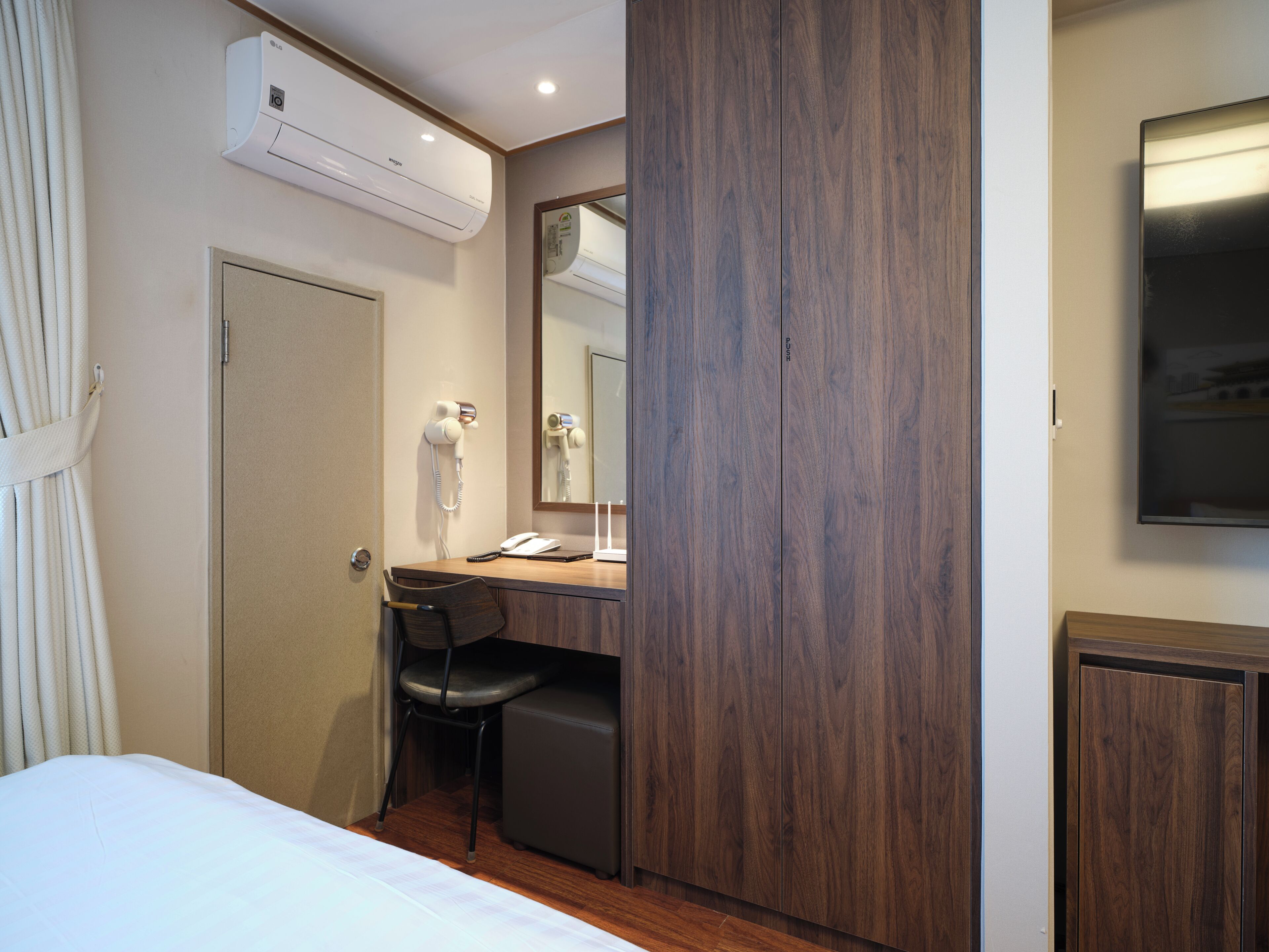 A hotel room with a bed, a desk with a chair, a hairdryer, a mirror, and an air conditioning unit.