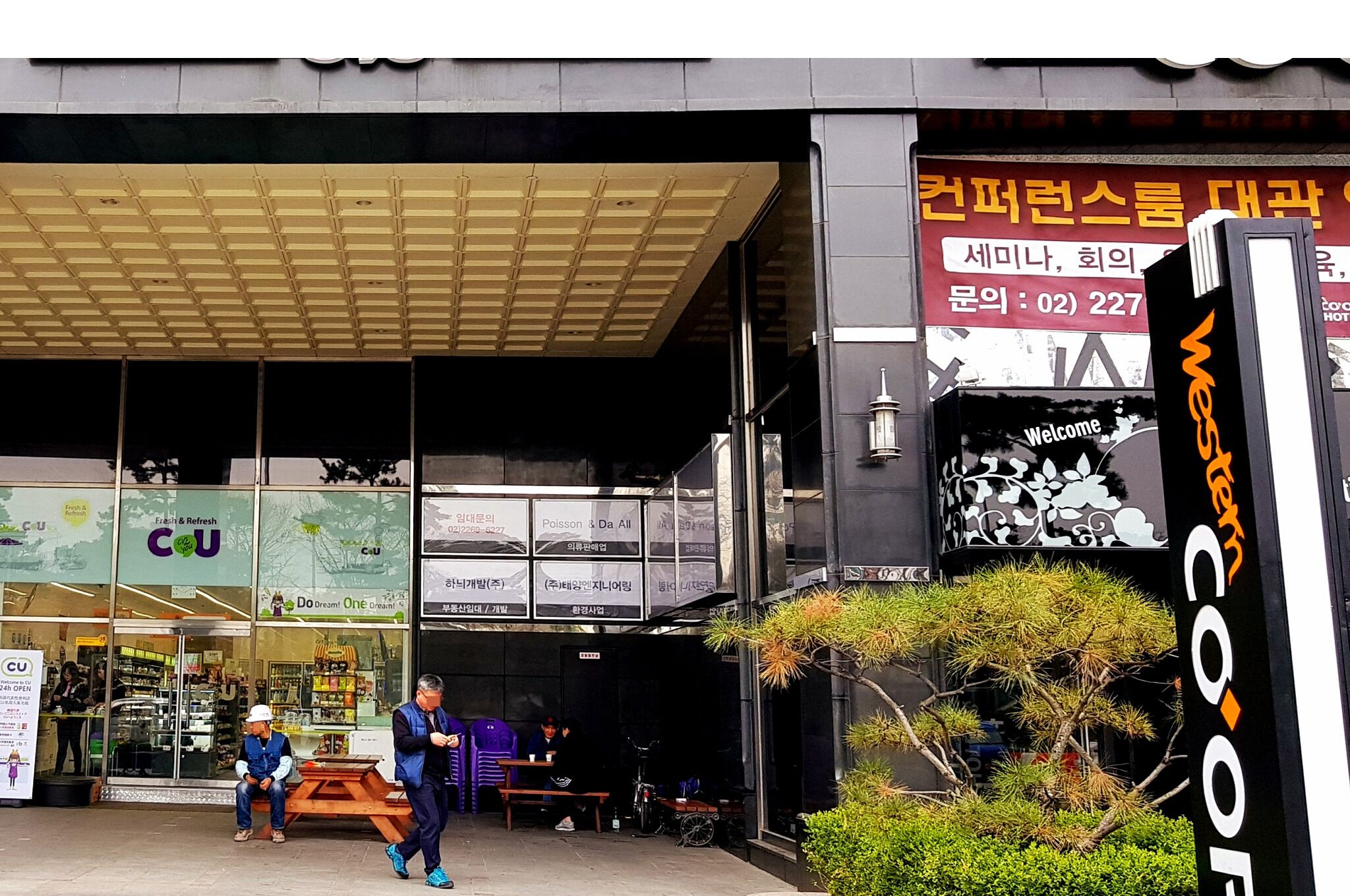 Ein Hotel mit Eingangsbereich und überdachter Außenbereich zum Sitzen, einem Schild mit koreanischer Schrift und einem 'Welcome'-Schild.