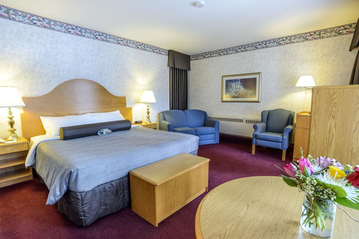 A hotel room with a bed, a desk, a chair, a small table, and a vase with flowers.