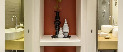 A modern bathroom with a white sink, a mirror, a black handbag on a shelf, and a vase with plants.
