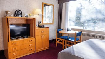 Ein Hotelzimmer mit einem Holz-Fernsehschrank, einem Flachbildfernseher, einem kleinen Tisch mit einem Stuhl, einem Fenster mit Blick auf Bäume und einem Spiegel an der Wand.