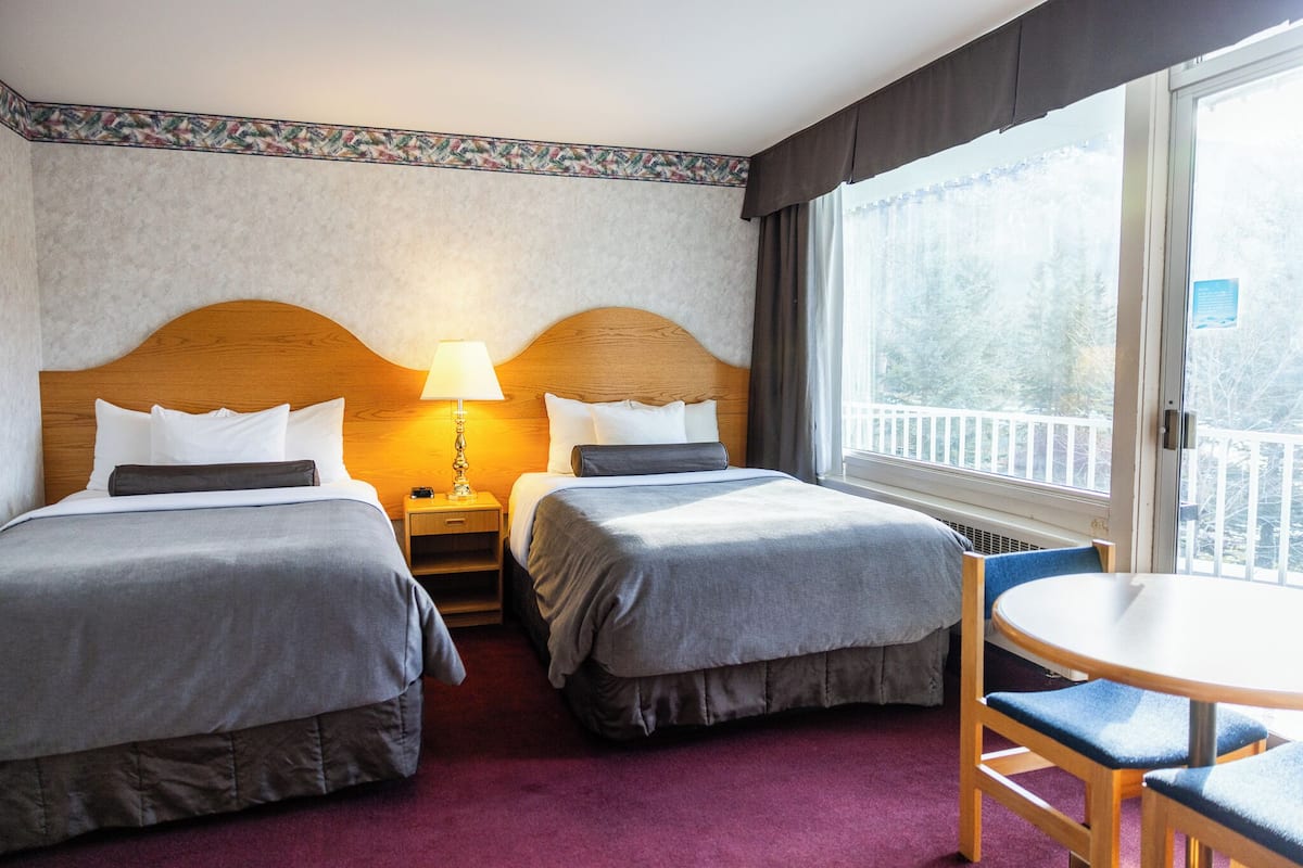 A hotel room with two beds, a round table, and a view of trees through the window.