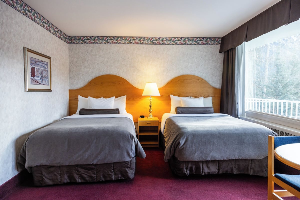A hotel room with two beds, a nightstand with a lamp, a framed picture on the wall, and a window with a view of trees.