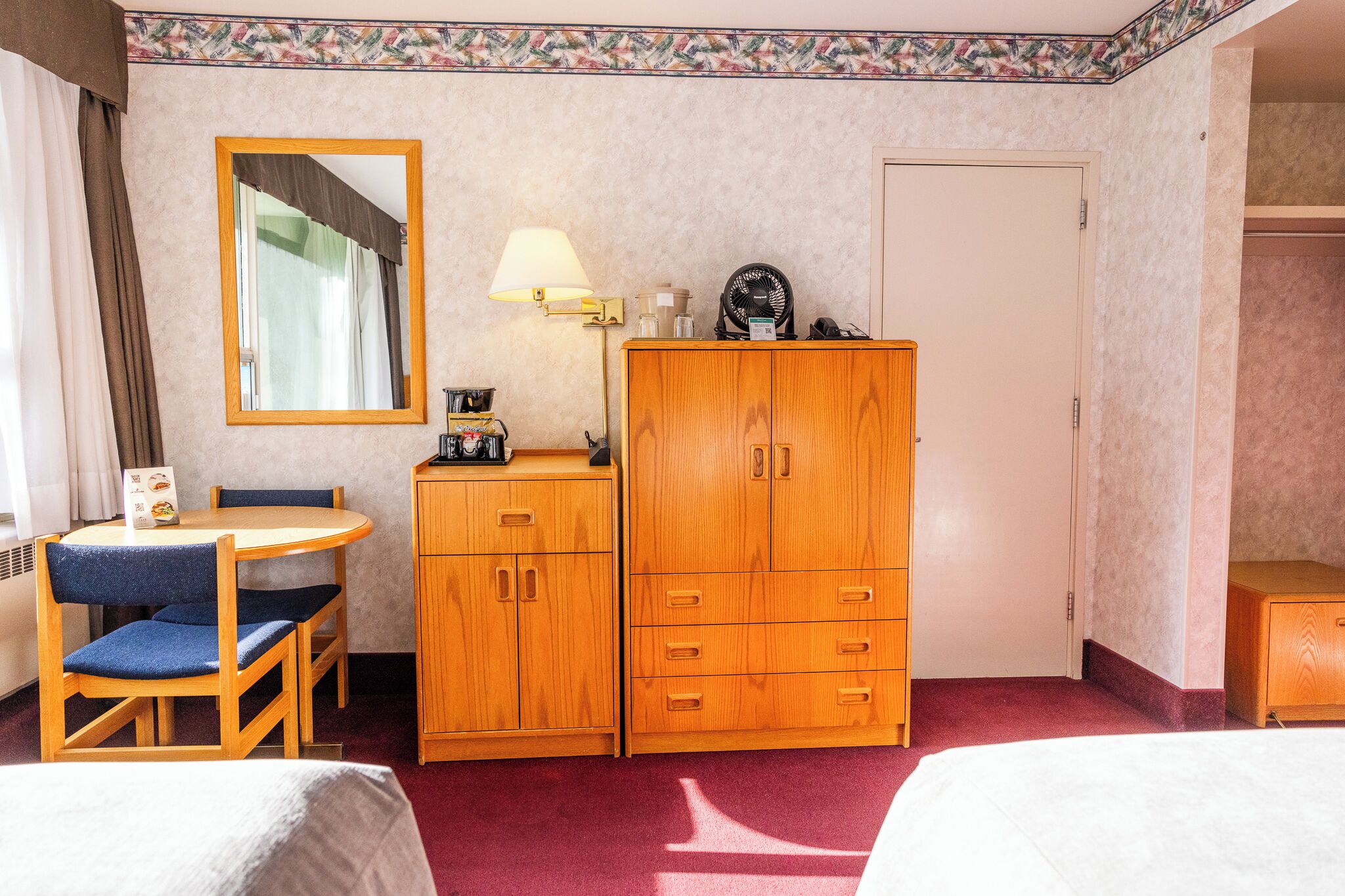 A hotel room with a wooden desk, two chairs, a wardrobe, a mirror, and a lamp.