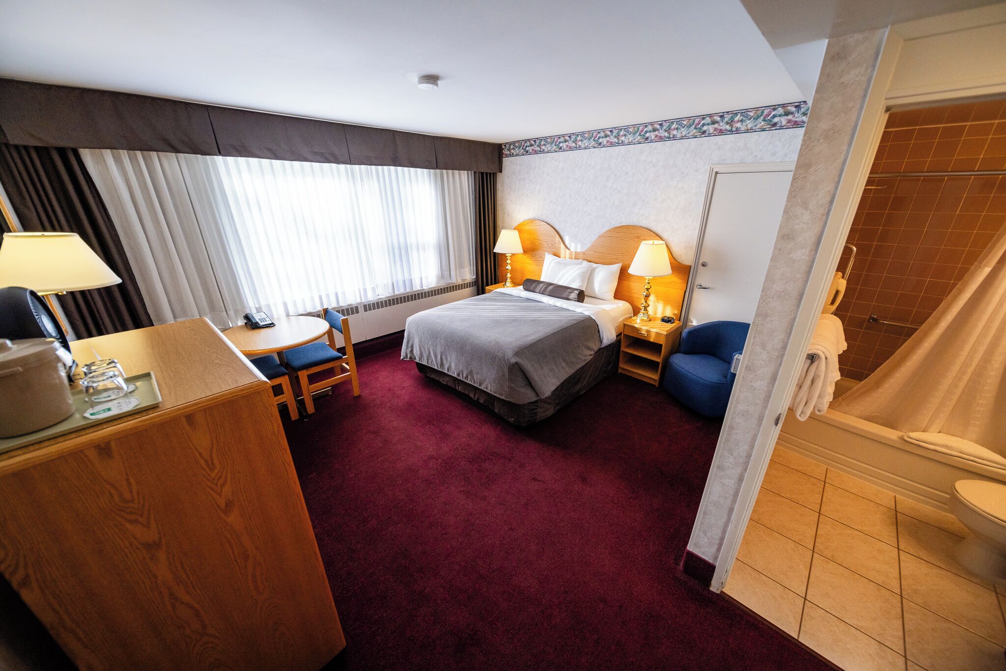 A hotel room with a bed, bedside tables, a desk, a chair, a window with curtains, and a bathroom with a shower and toilet.