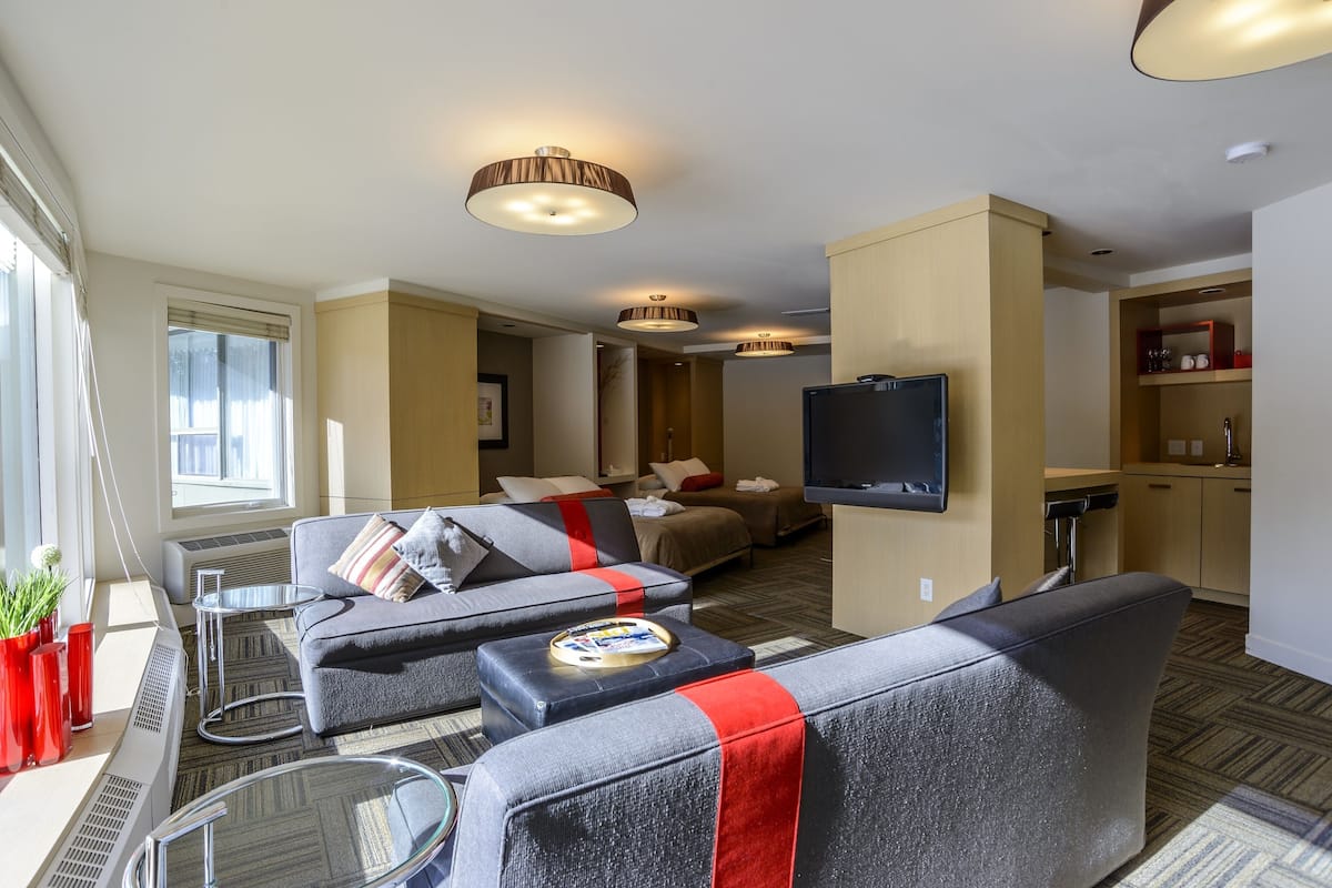 A modern hotel room with a sofa set, a flat-screen TV, a small kitchen area, and a dining table.