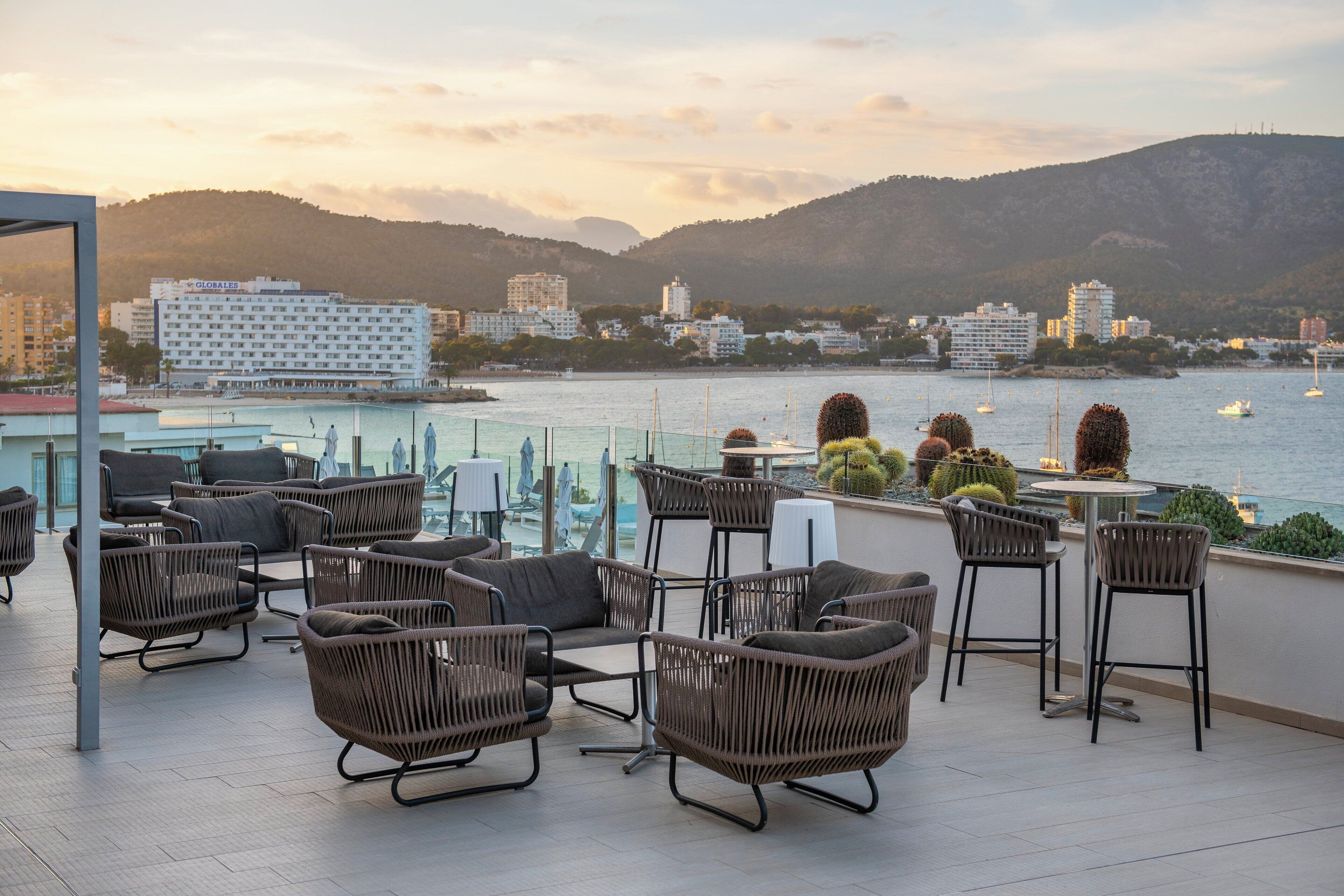 A rooftop terrace with wicker furniture, a pool, and a view of the city and hills.
