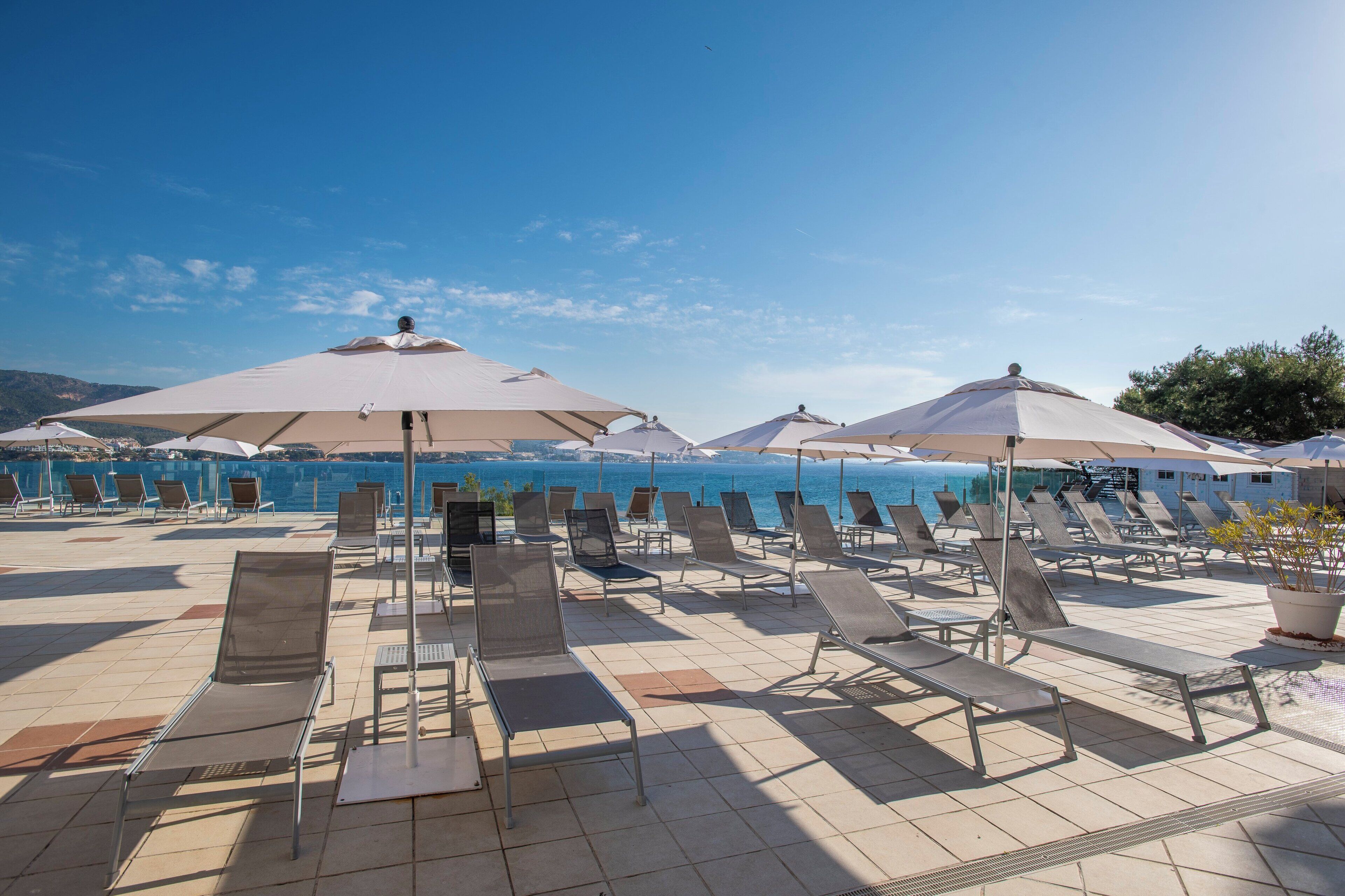 Een zonnig terras buiten met lounge fauteuils en parasols, met uitzicht op een waterlichaam.