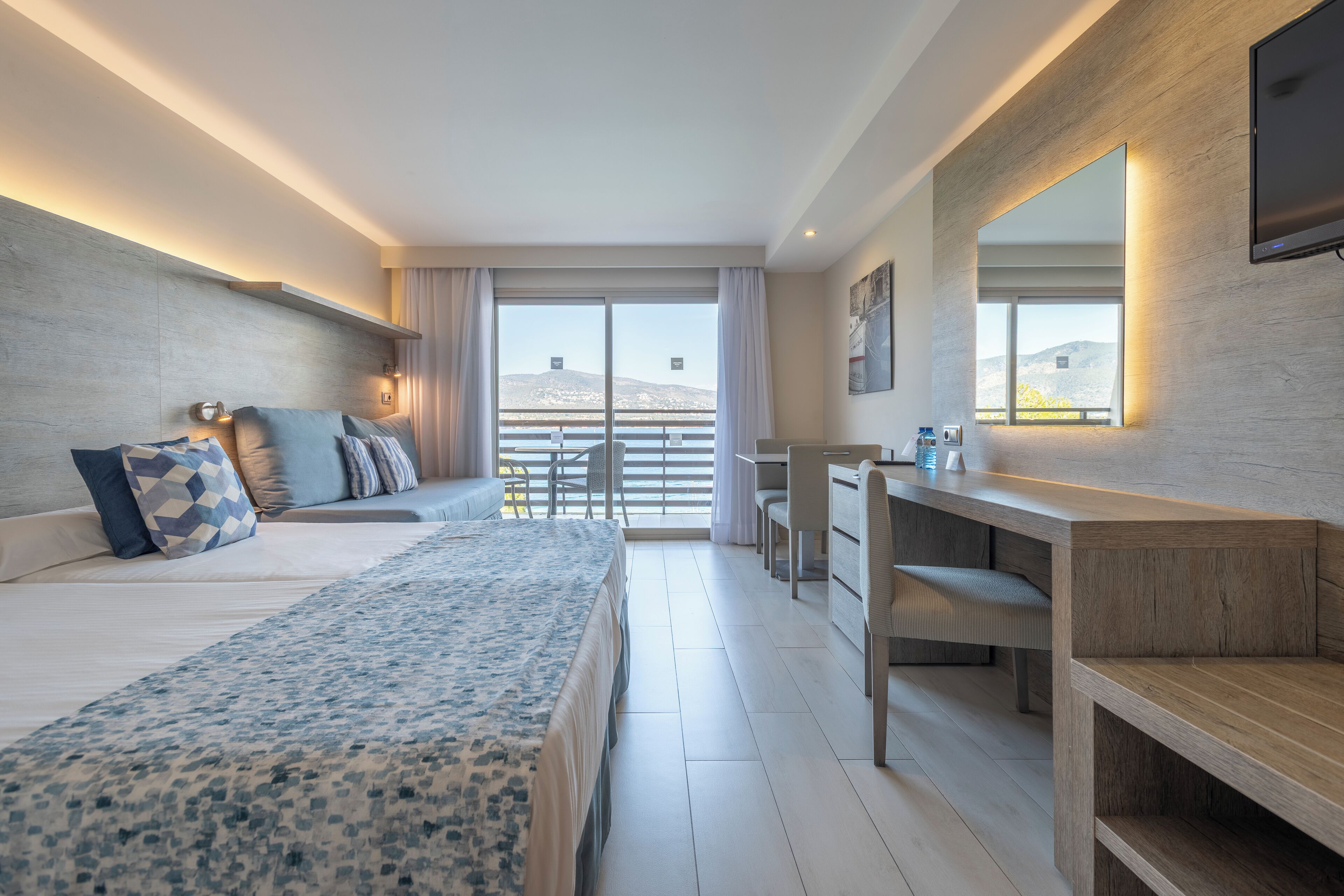 A modern hotel room with a large bed, a desk, and a balcony with a view of the mountains.