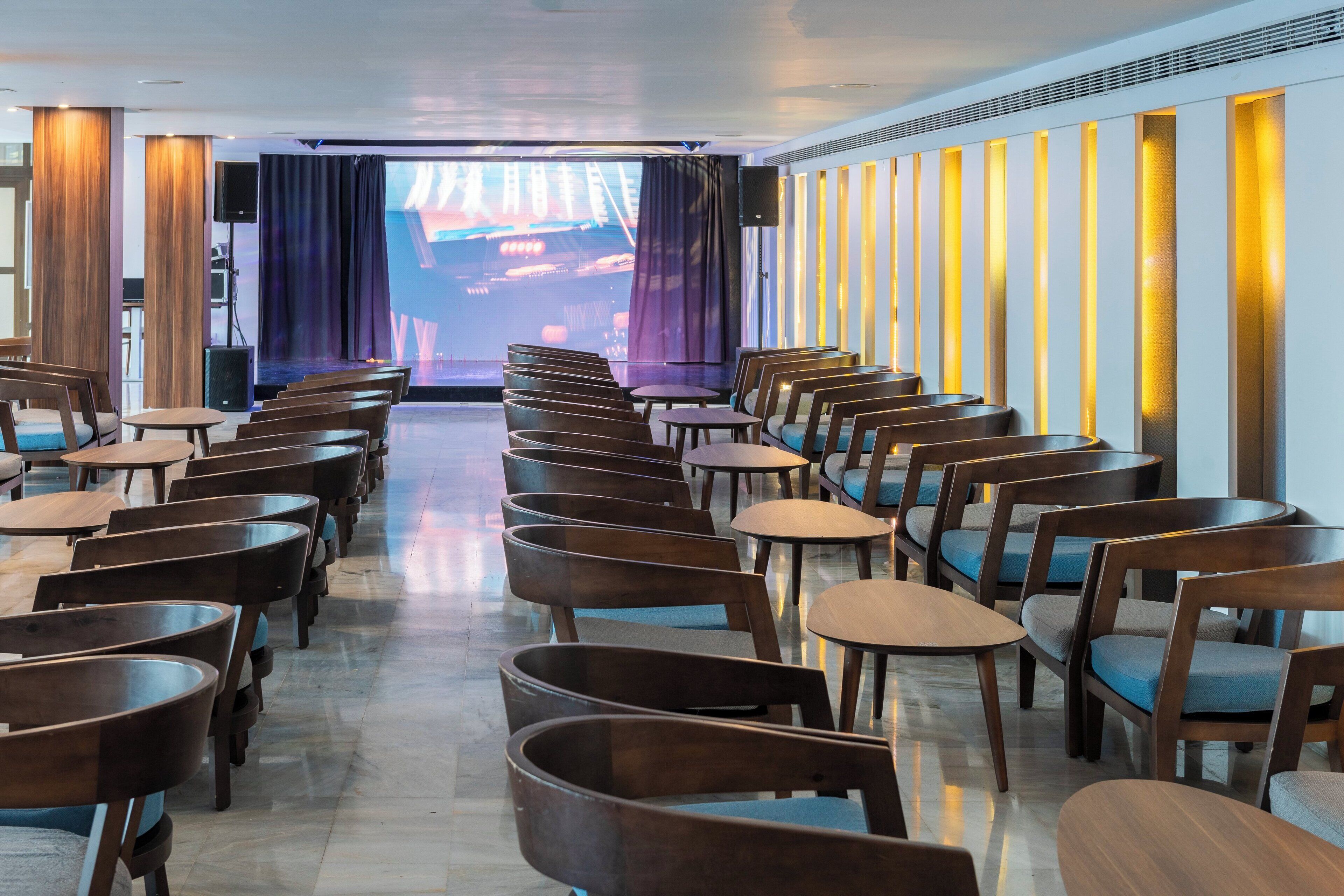 Un salón de conferencias con filas de sillas y mesas de madera, una pantalla grande al frente y un orador a la izquierda.
