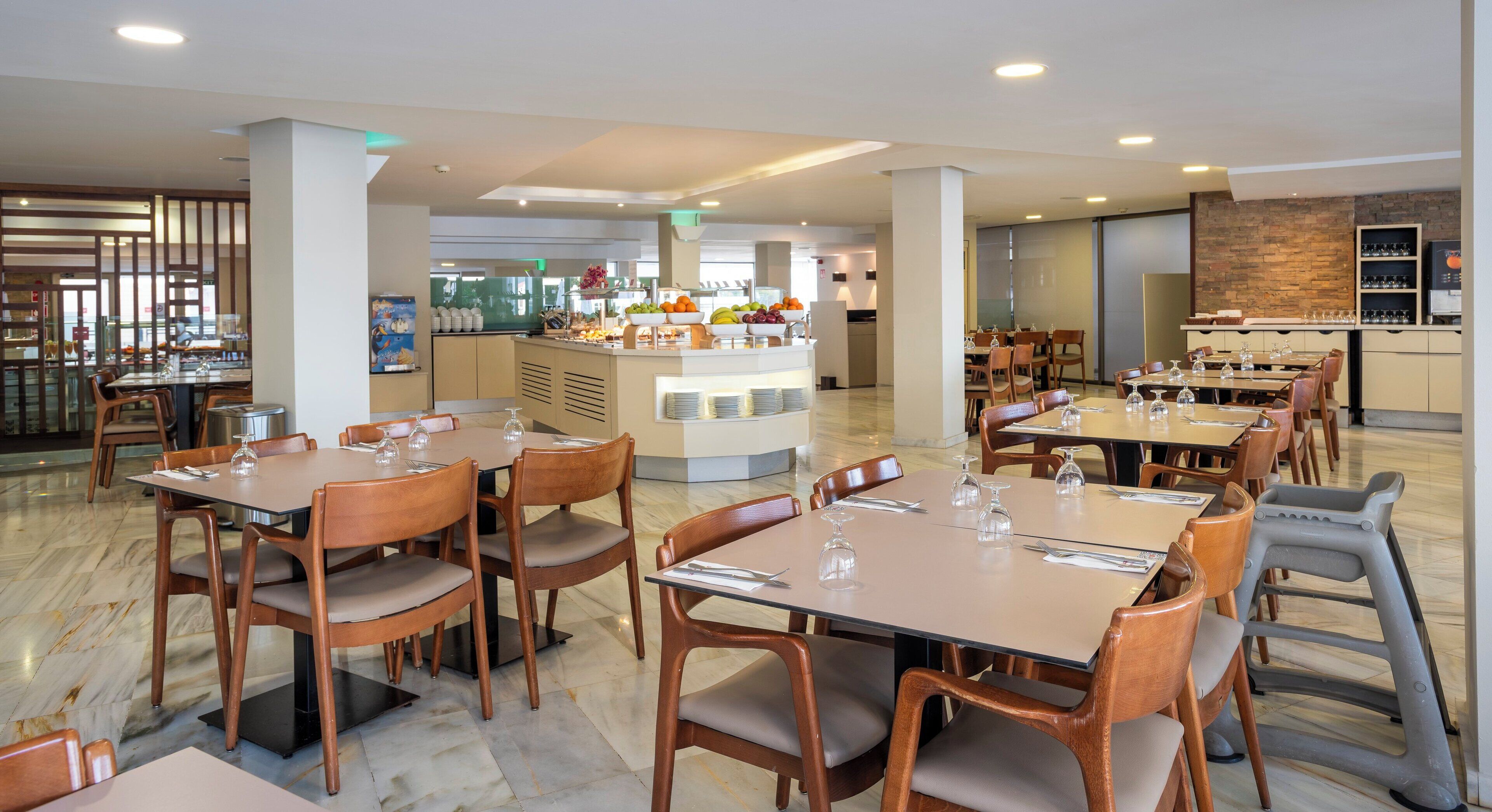 Un vaste espace de restauration d’hôtel, avec de nombreuses tables et chaises, un buffet central proposant une grande variété de plats, et une cuisine située à l’arrière-plan.