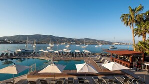 Zona de piscina amb gandules i ombrel·les, amb vista a una marina amb vaixells i una serralada de muntanyes a la distància.