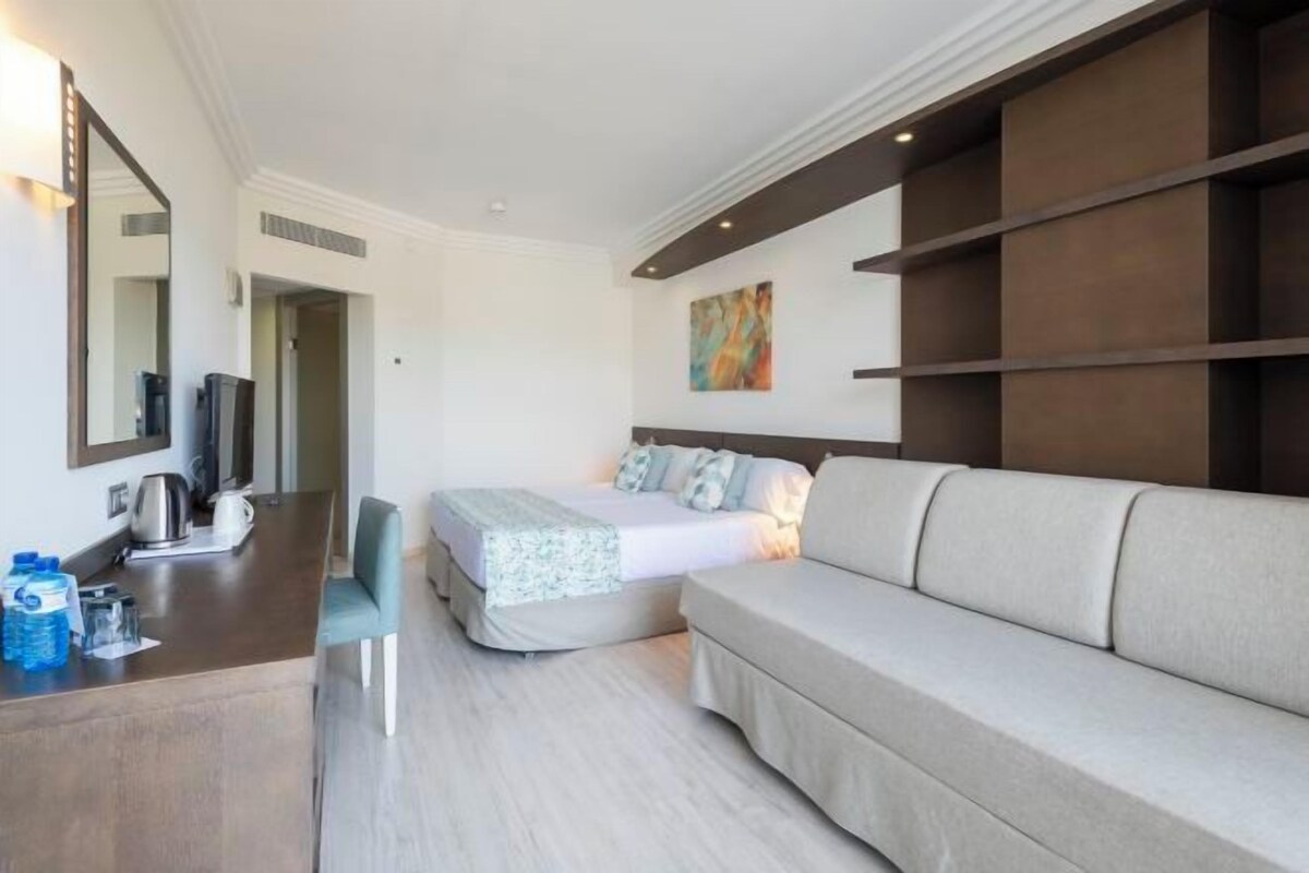 A hotel room with a bed, a sofa, a desk with a chair, a television, and a mirror.