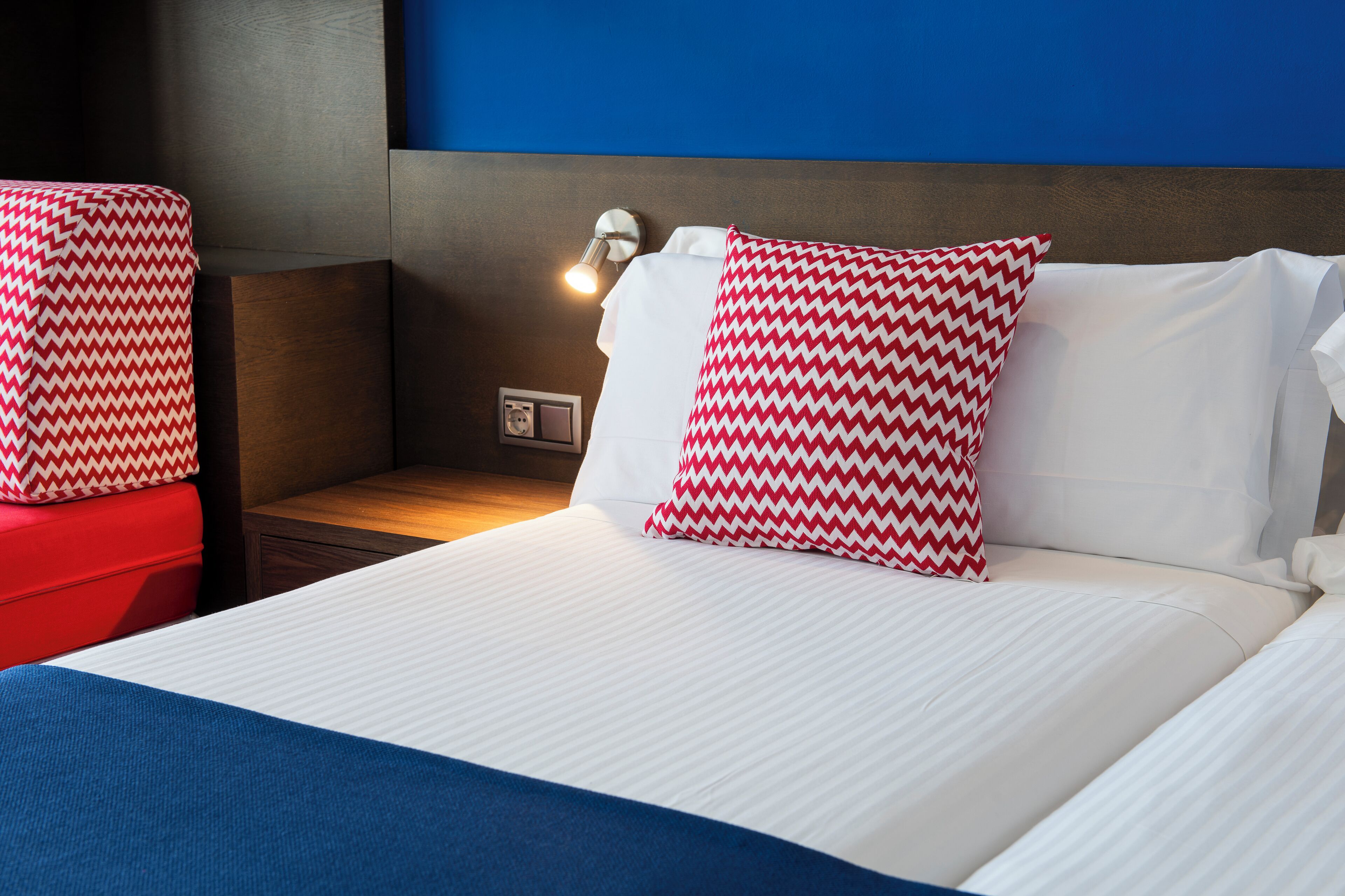 A hotel room with a bed, a nightstand, a red chair, and a wall-mounted lamp.