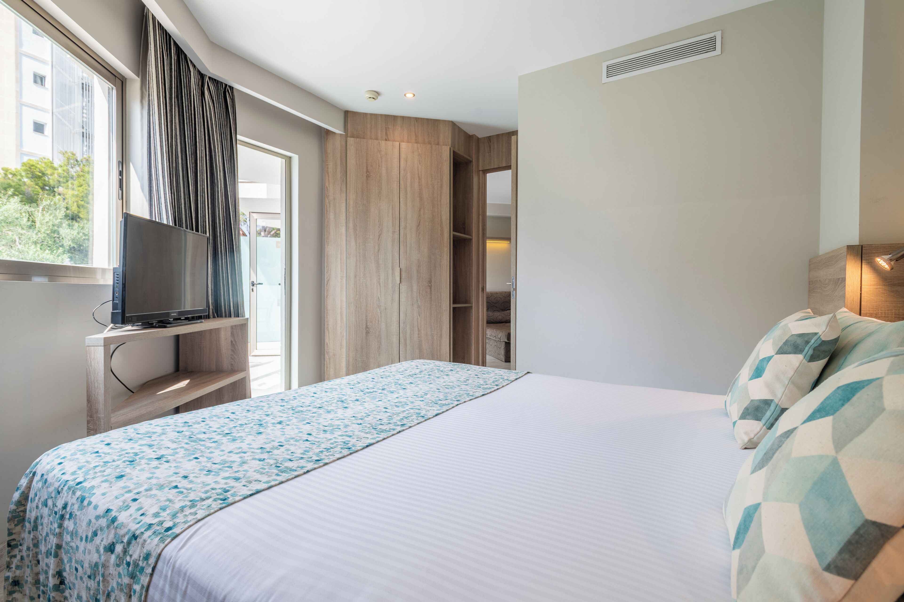 A hotel room with a bed, a TV on a wooden stand, a wardrobe, and a window with curtains.