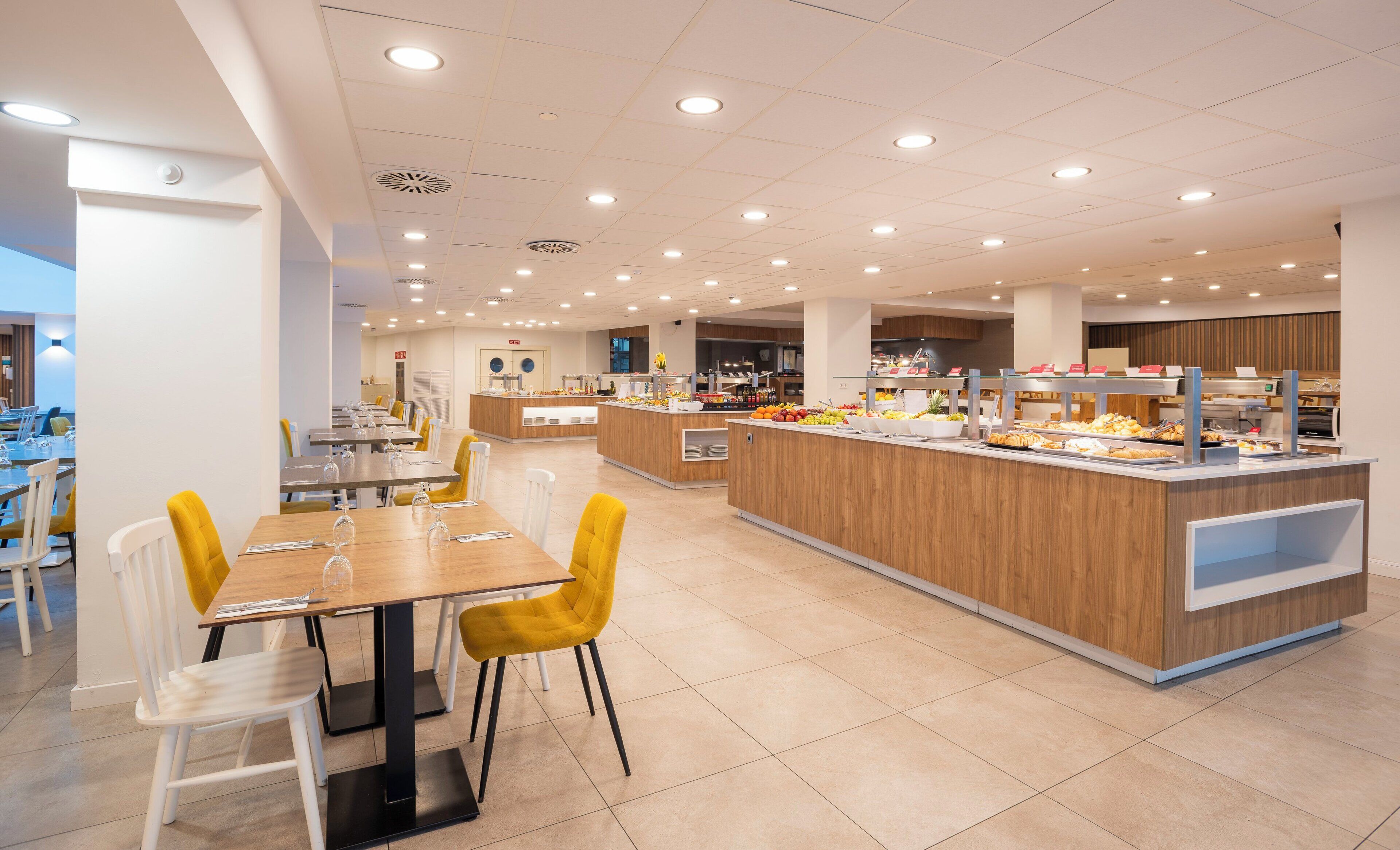 Een ruime cafetaria met meerdere eettafels en stoelen, een centraal buffetgedeelte met diverse gerechten, en een schone, goed verlichte omgeving.