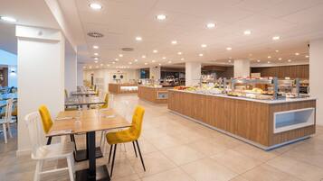 Een ruime cafetaria met meerdere eettafels en stoelen, een centraal buffetgedeelte met diverse gerechten, en een schone, goed verlichte omgeving.
