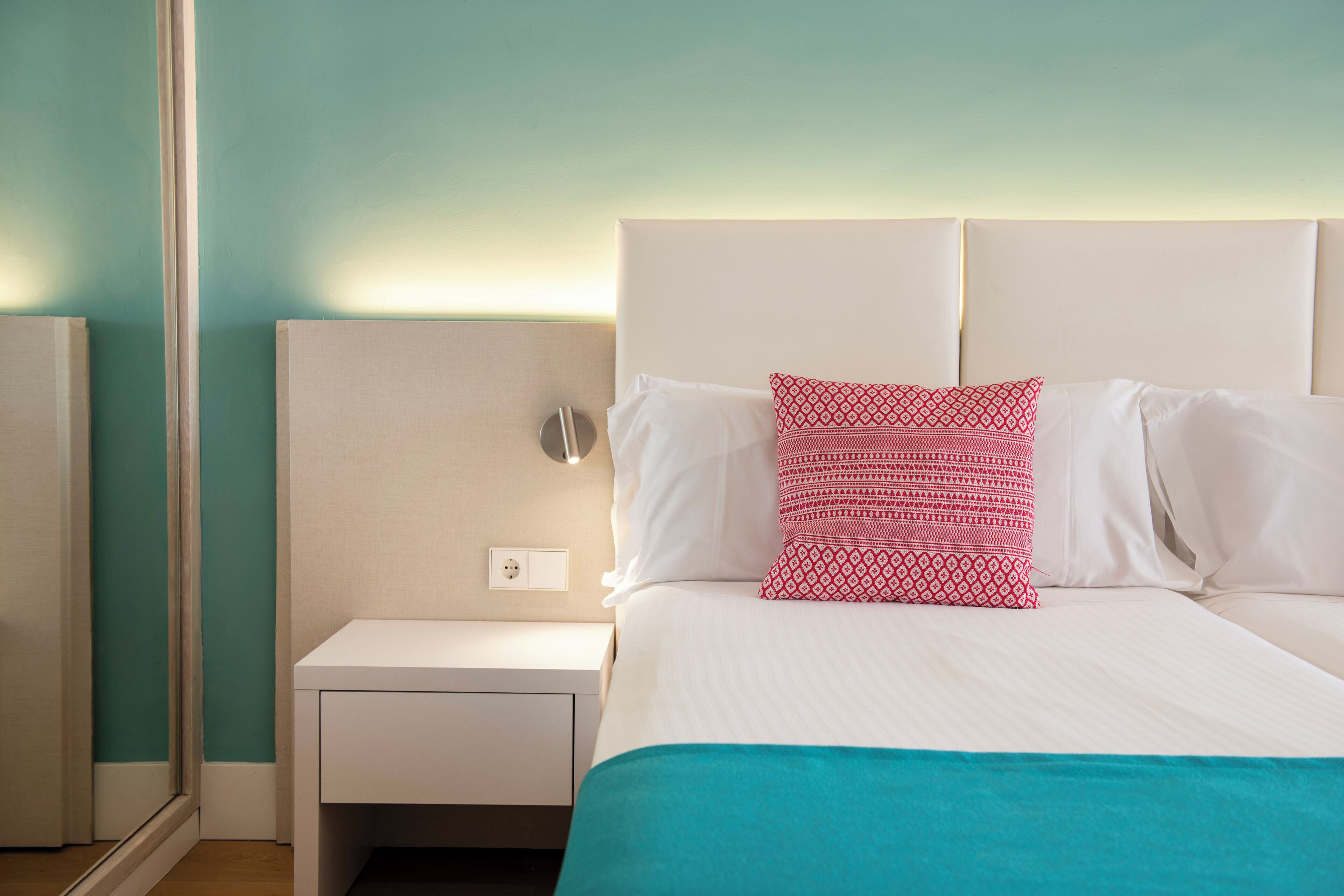 Una cama bien hecha con ropa blanca y un plaid verde azulado, una mesita de noche blanca con una lámpara, y un espejo en la pared.