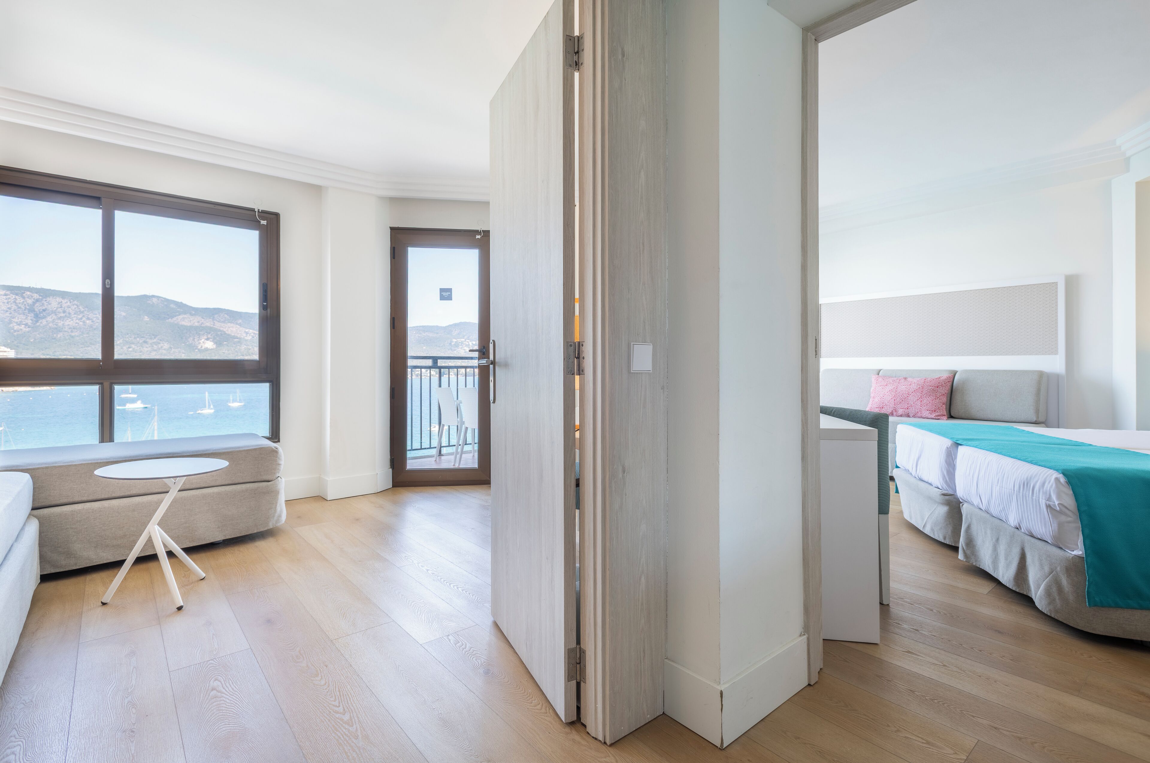 Et moderne hotelværelse med et stort vindue, en balkon, en seng med turkis sengetæppe og et siddeområde med et rundt bord.