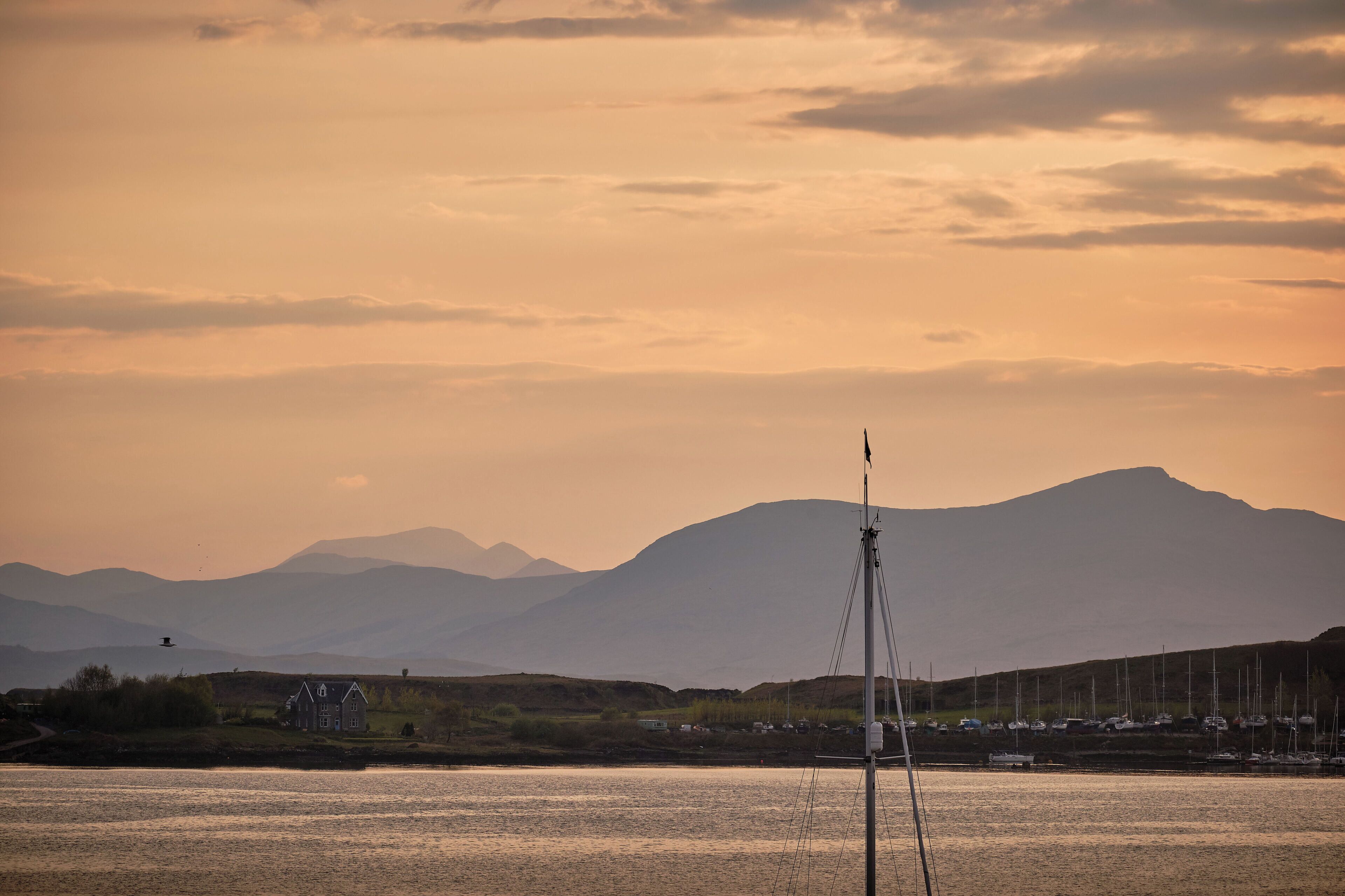 A sailboat on calm waters with a mountainous backdrop and a small building on the shore.