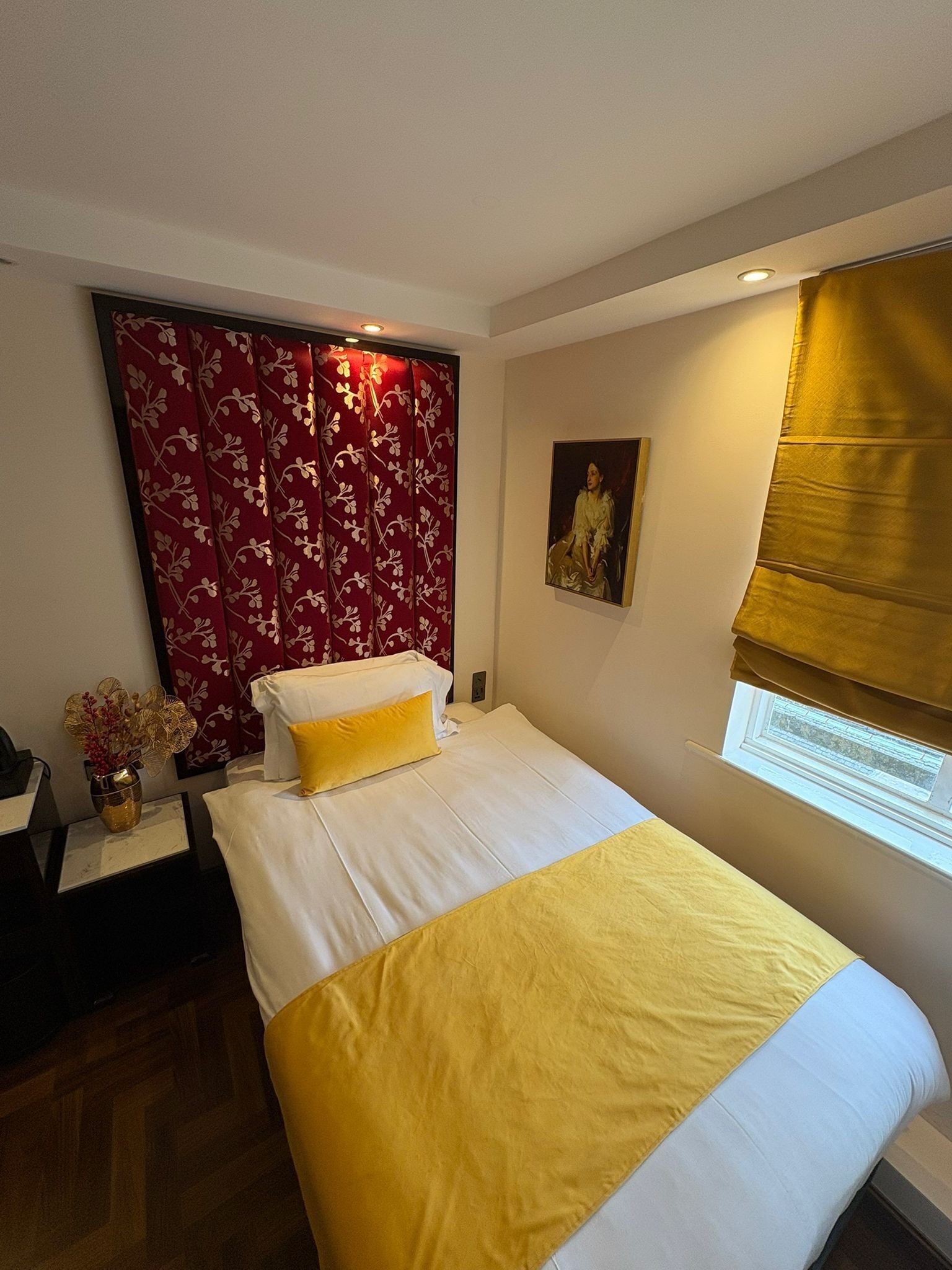 A hotel room with a bed, a window with a blind, a painting on the wall, and a decorative vase on a nightstand.