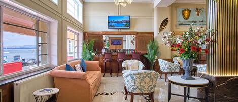 A spacious lobby with a large window offering a view of the sea, a comfortable seating area with a sofa and armchairs, a bar area with a television, and decorative elements like a shield and antlers on the wall.
