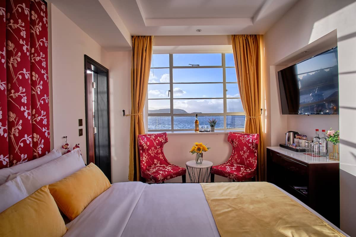 A hotel room with a bed, two red floral chairs, a small table with a vase of flowers, a TV on the wall, and a window with a view of the sea.