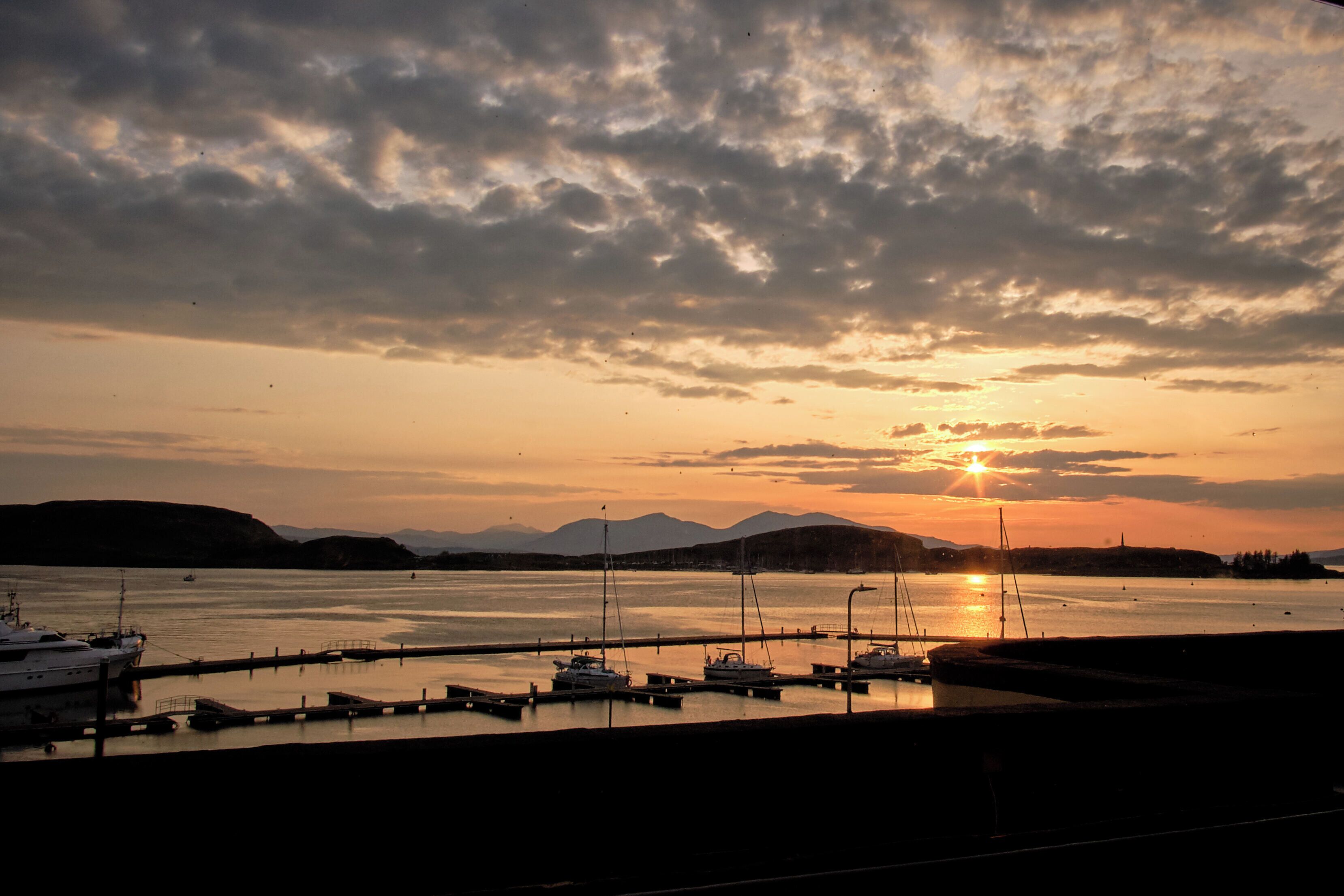 Ein Yachthafen mit vertäuten Booten, ein Sonnenuntergang im Hintergrund und Berge in der Ferne.